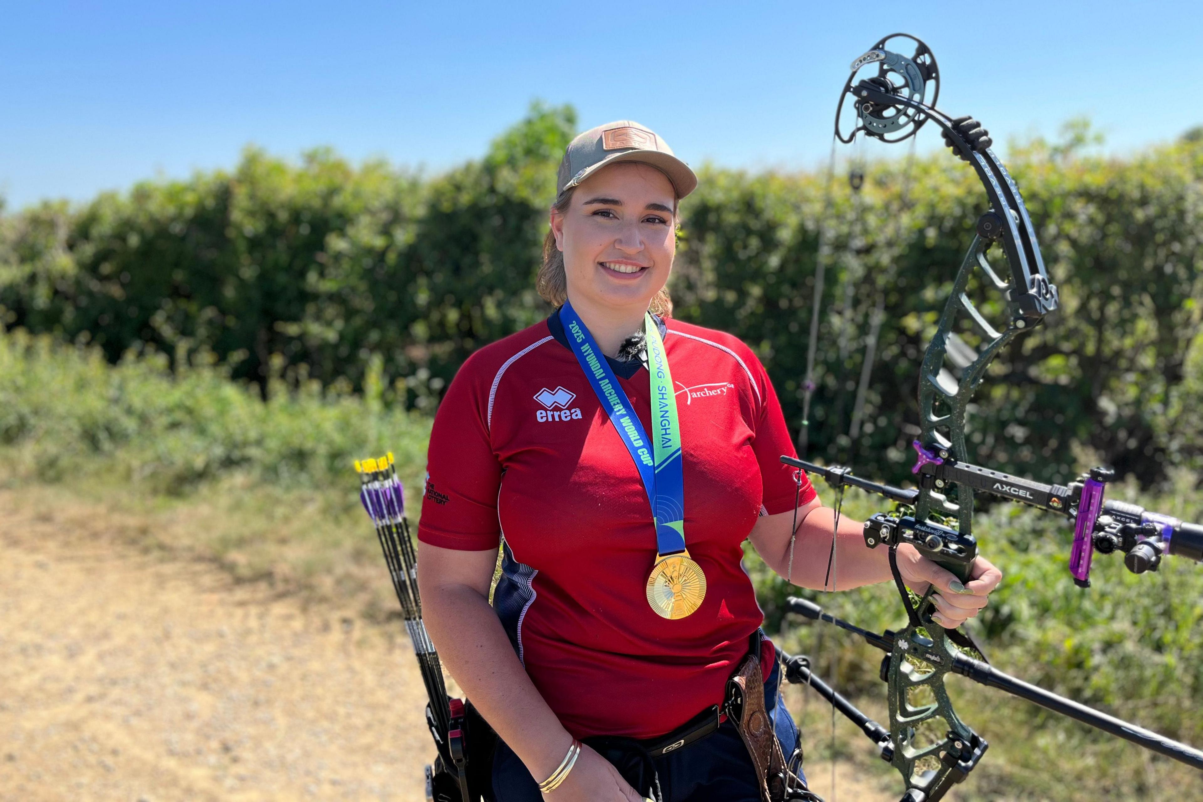 A woman is donning a red sporty shirt and wearing a gold medal around her neck, and is looking straight at the camera smiling while holding a bow that looks very sophisticated.