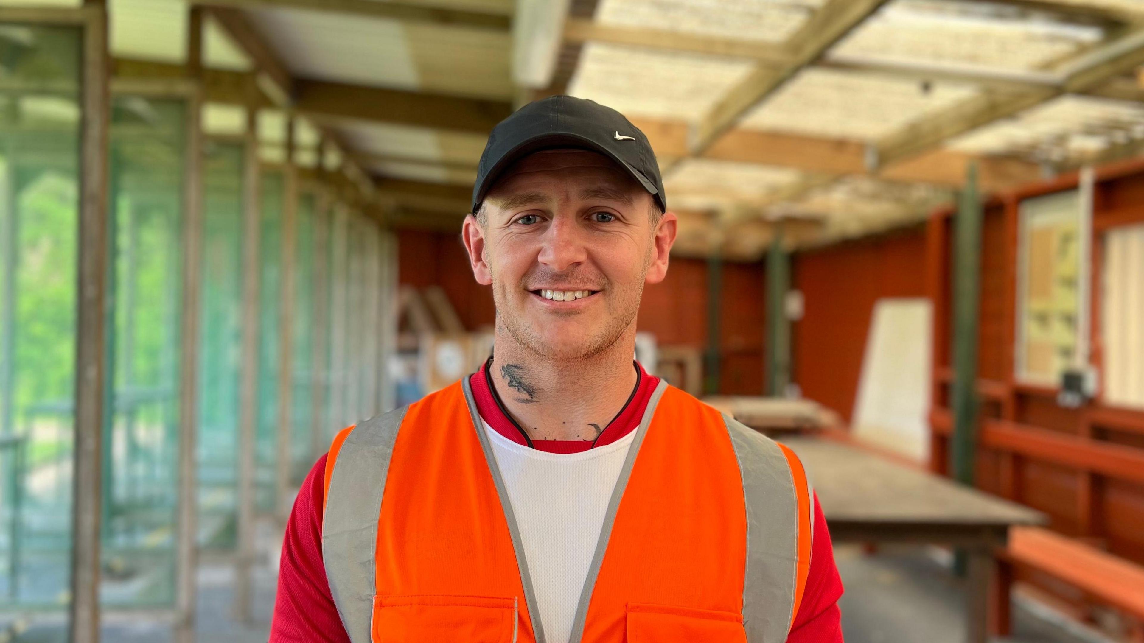 Ben Videgrain is standing in a shooting range. He is smiling at the camera and is wearing a black baseball cap, a white and red t-shirt and an orange high viz vest over it. He has a tattoo on the left side of his neck.
