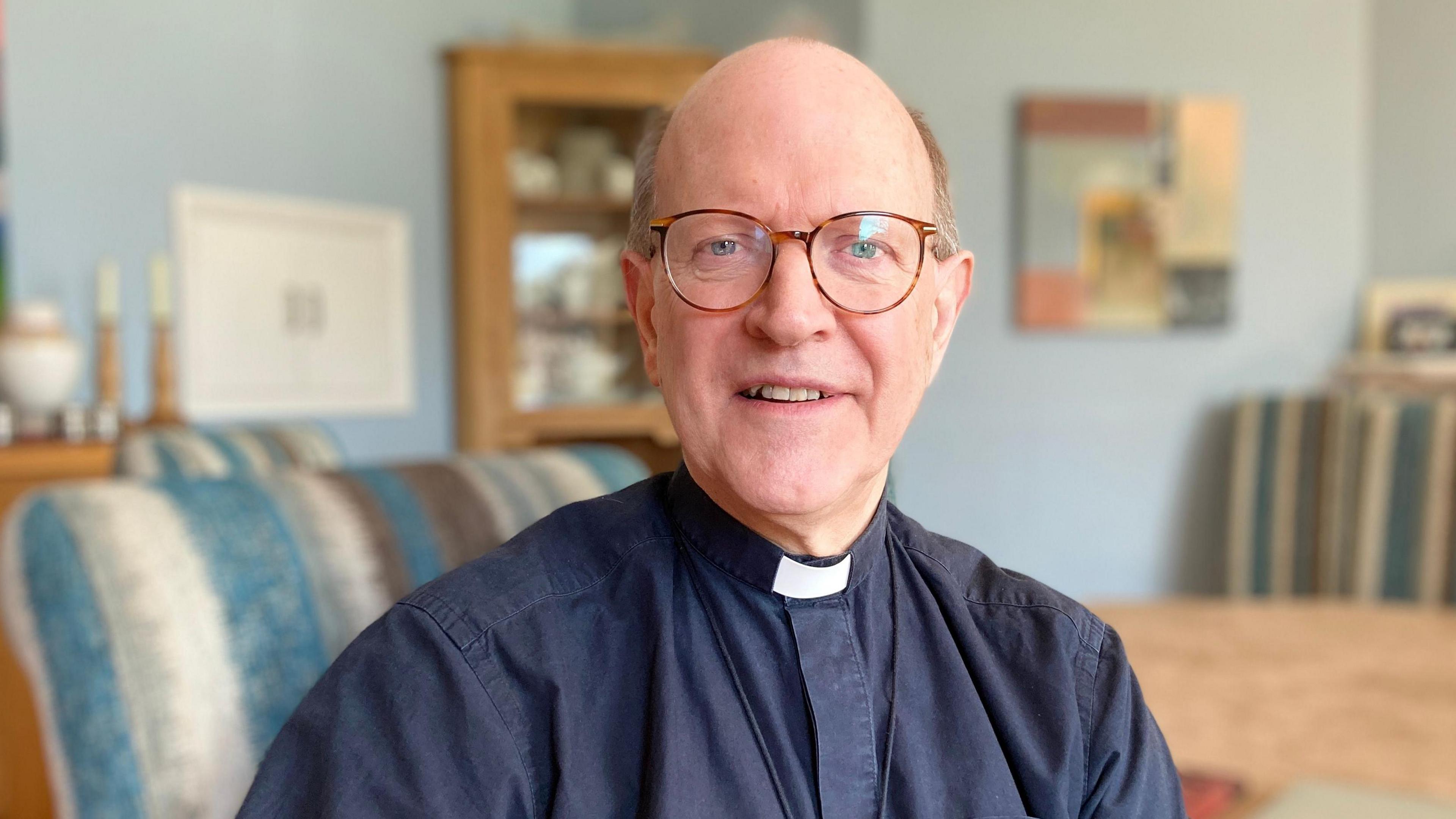 A close-up image of The Bishop of St Edmundsbury and Ipswich. He is sitting in a chair and wearing glasses, a clerical collar and a  black top.