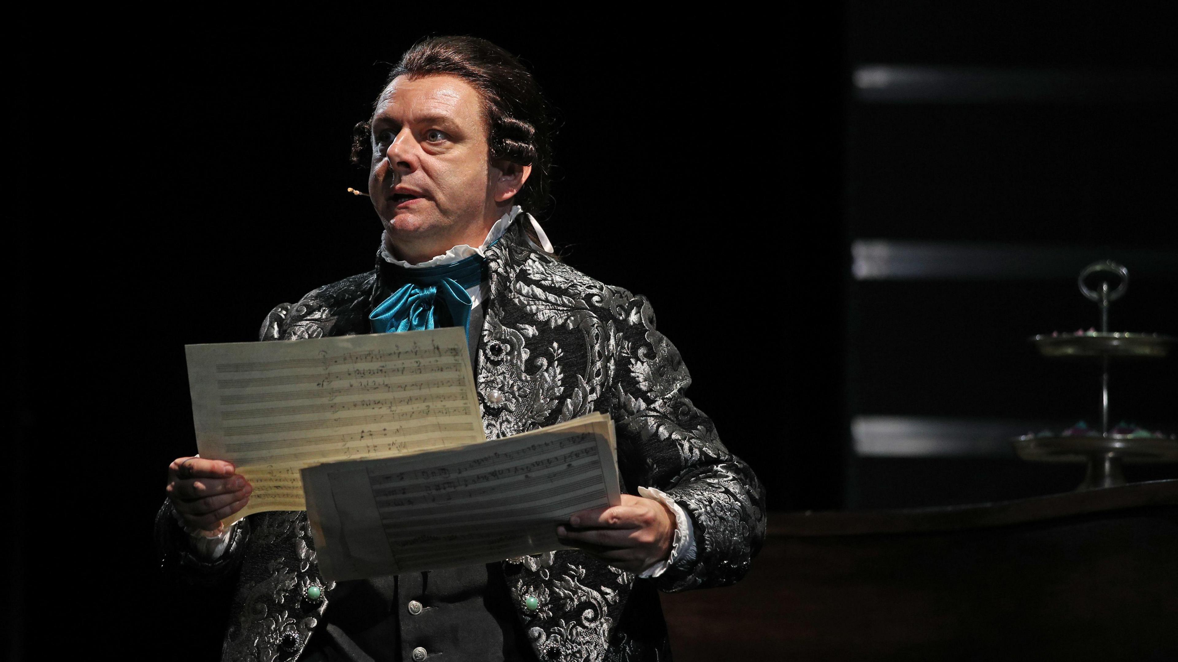 A picture of Michael Sheen playing the role of Antonio Salieri during a media call for Amadeus at Sydney Opera House on December 30, 2022 in Sydney, Australia. He is wearing a patterned sliver and black jacket and is holding papers of music. The spotlight is on him. 