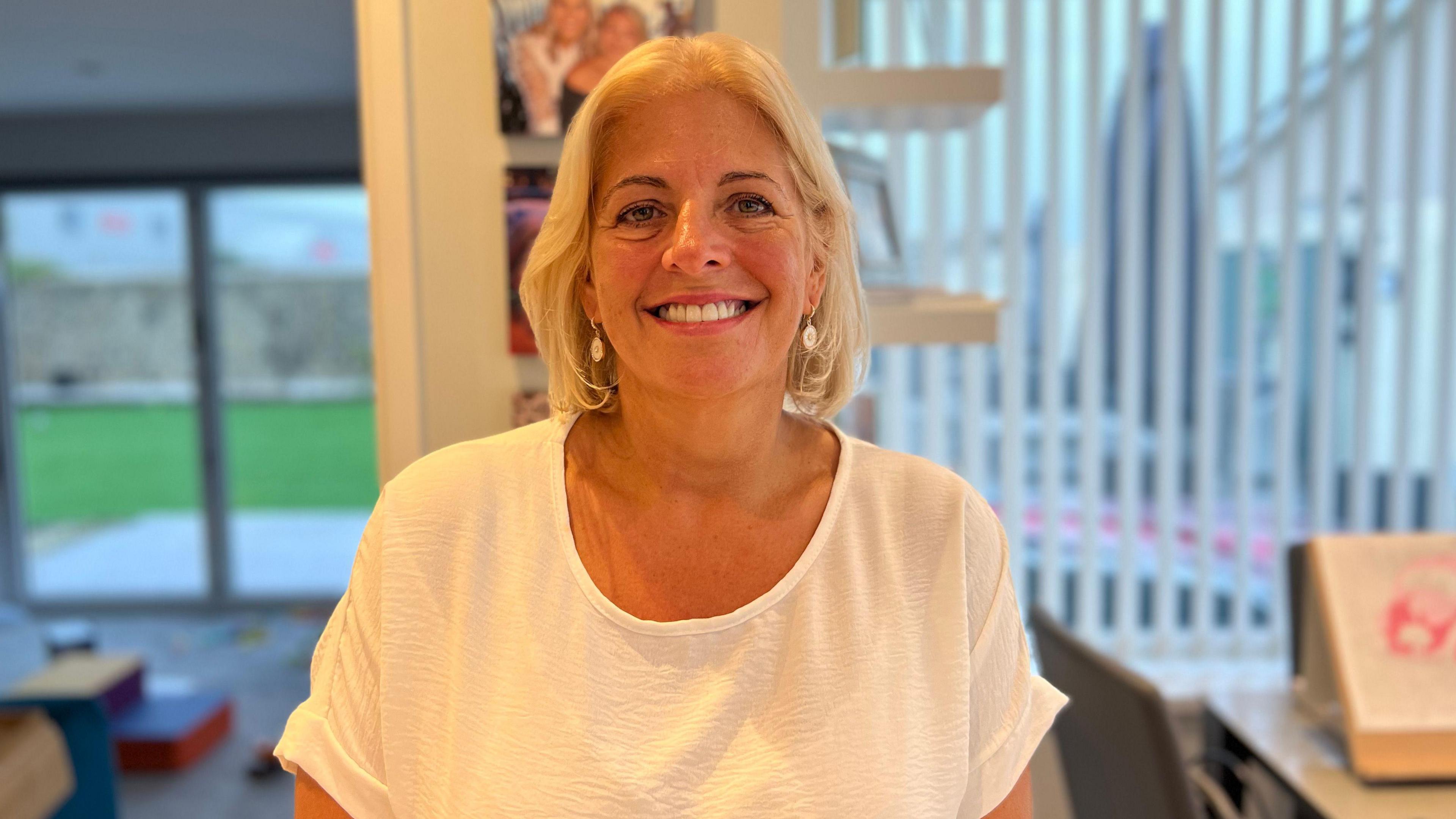 Photo of a woman with short blonde hair smiling at the camera. She is wearing a white T-shirt with silver earrings.