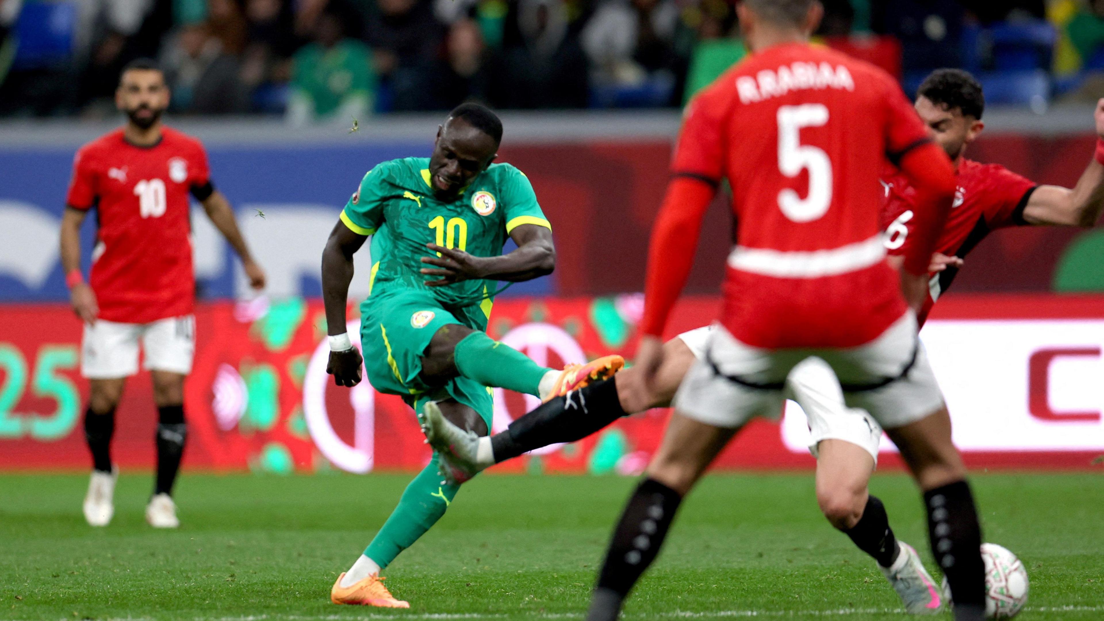 Sadio Mane in all-green kit is seen after taking a right-footed shot in a game between Senegal and Egypt at the 2025 Africa Cup of Nations. He has his head angled downwards with a strained look on his face as his right leg extends up and out in taking the strike. Two Egyptian players in red jerseys, white shorts and black socks can be seen looking to block the strike, with one lunging across Mane with his right leg. Mohamed Salah can be seen looking on in the background