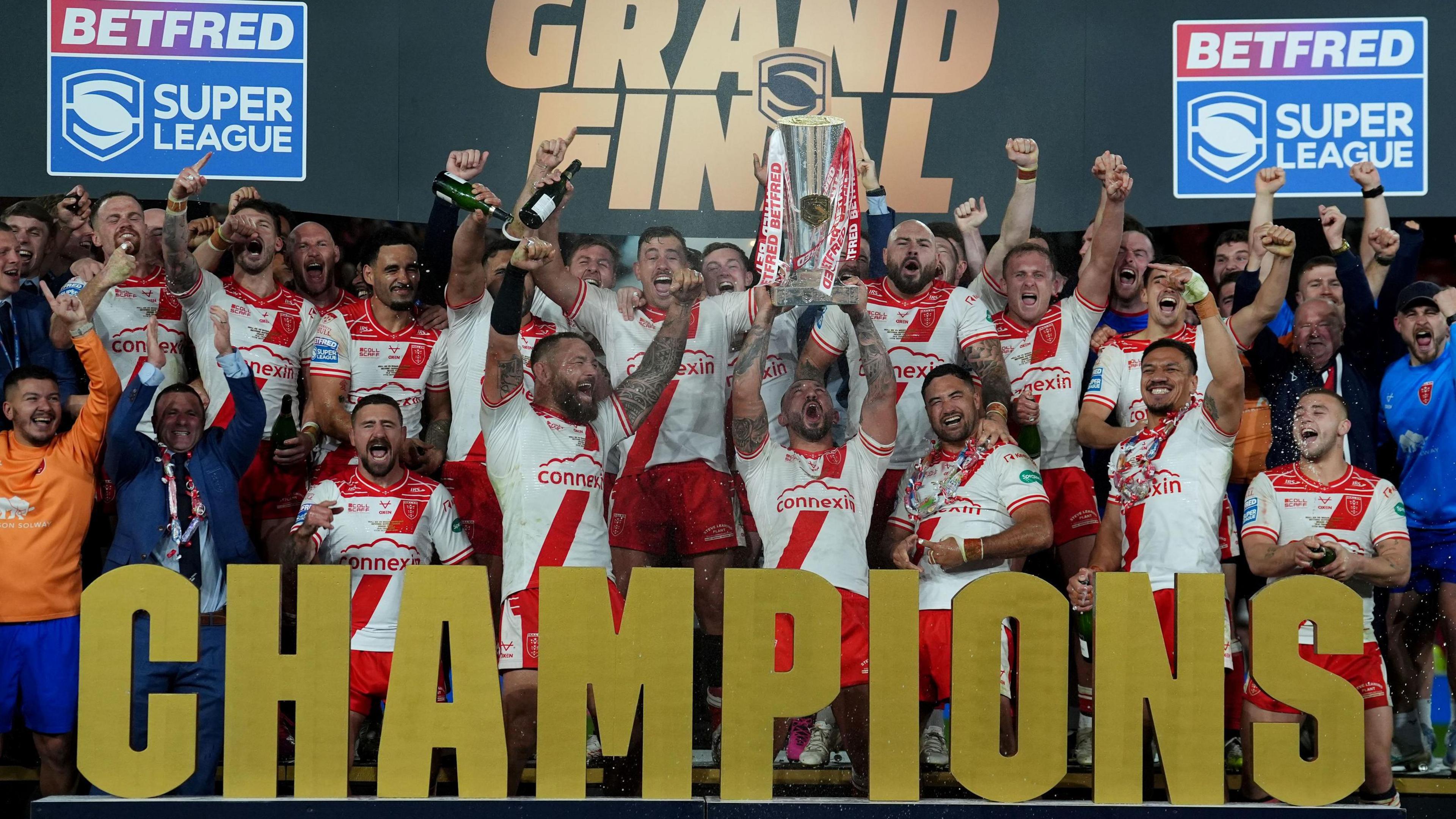 Hull KR players, wearing white shirts with a red stripe and red shorts celebrate with their arms in the air as the captain, standing in the middle, lifts a huge silver trophy into the air.