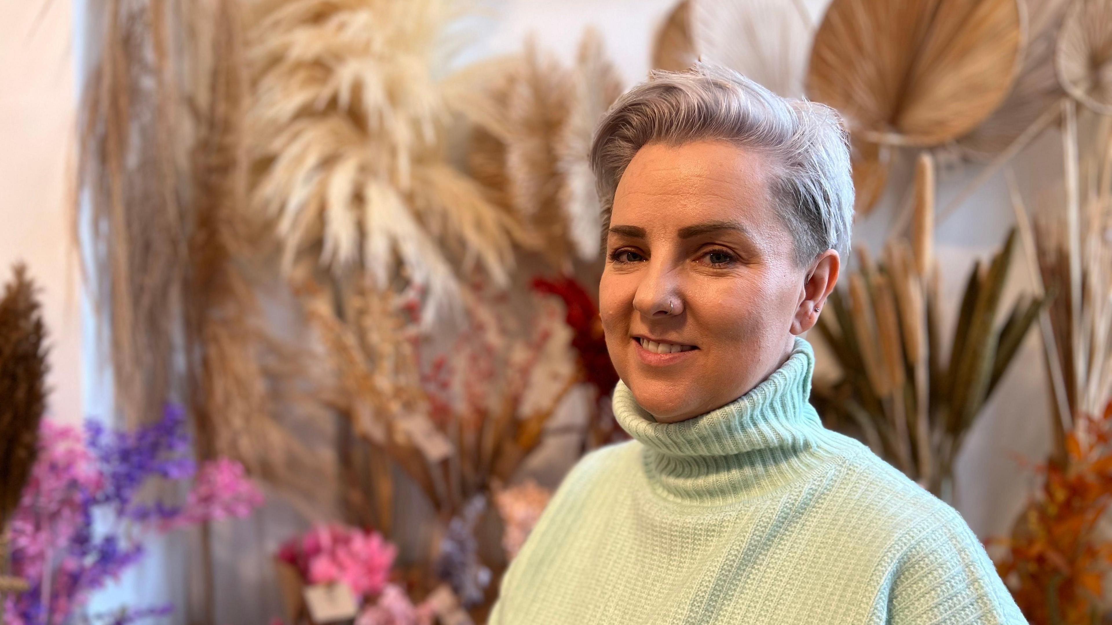 A woman with short dyed hair smiles and looks at the camera. She has a nose ring and is wearing a high-necked green jumper. In the background there is a colourful display of flowers for sale. 