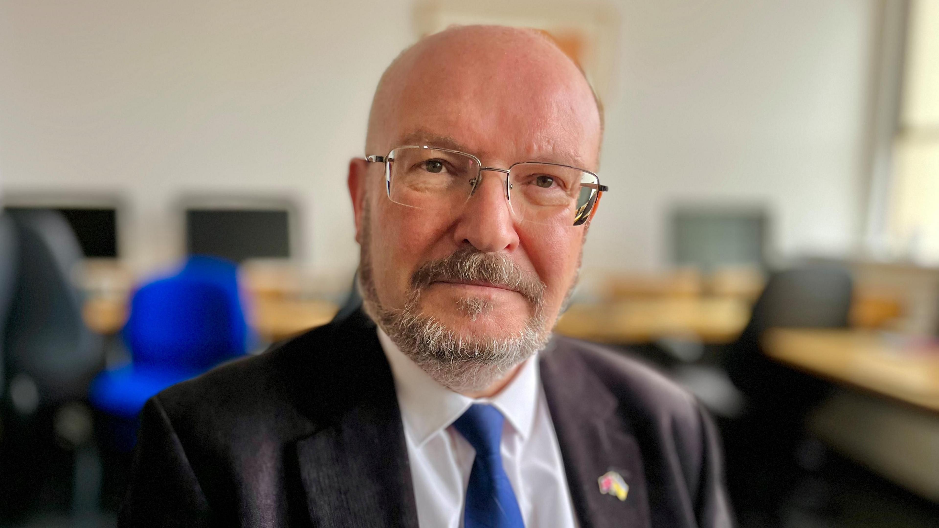 Graham Plant, a man who is sitting down inside a room with desks behind him. He is looking directly at the camera and smiling and is wearing a pair of glasses on his face, a black suit jacket, white shirt and blue tie.