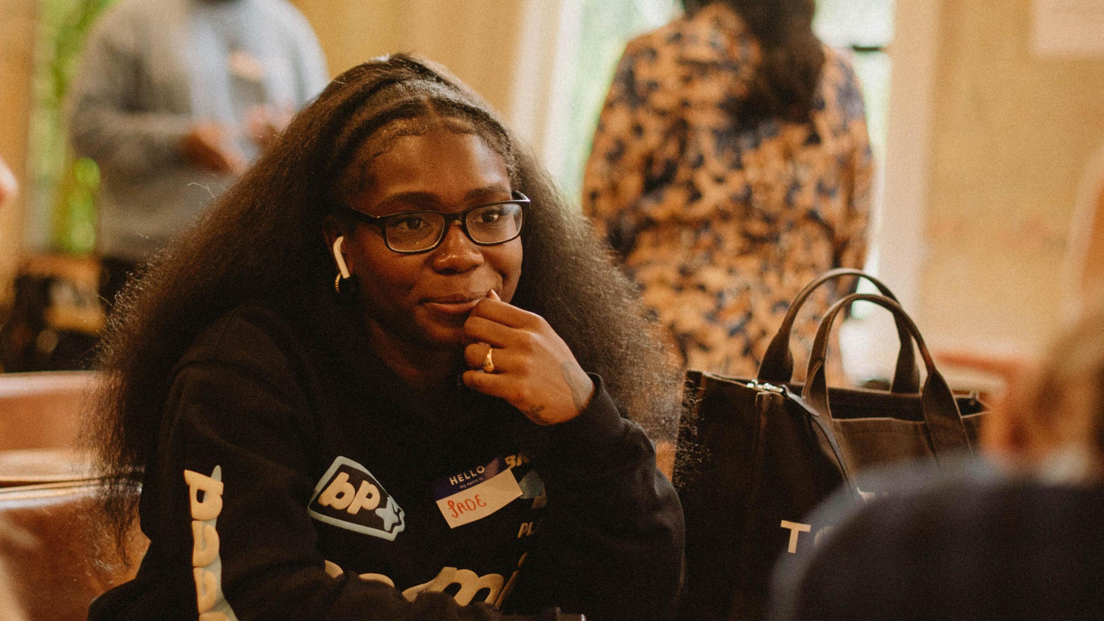 Jade Barnett has long brown hair and is wearing glasses and a black jukped with various logos on it. She is sitting at a table with one hand close to her face. There are a couple of people in the background.