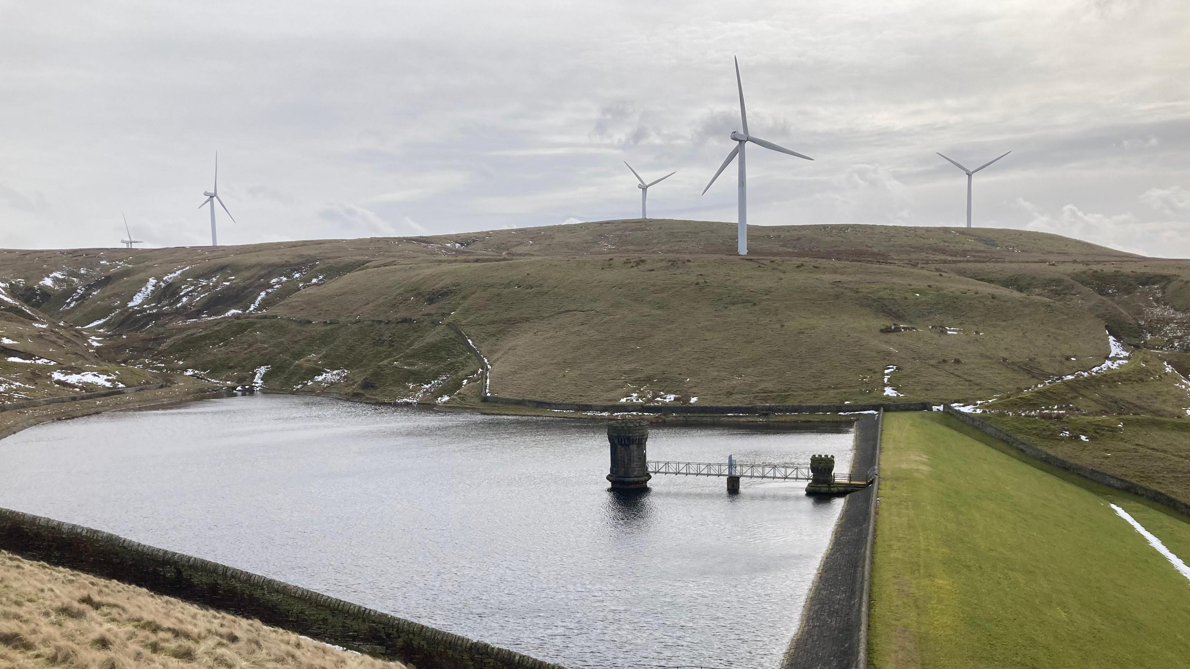 The wind farm of Scout Moor, with turbines seen on headland above a reservoir on an overcast day, with patches of snow seen in the clefts of the hillside.