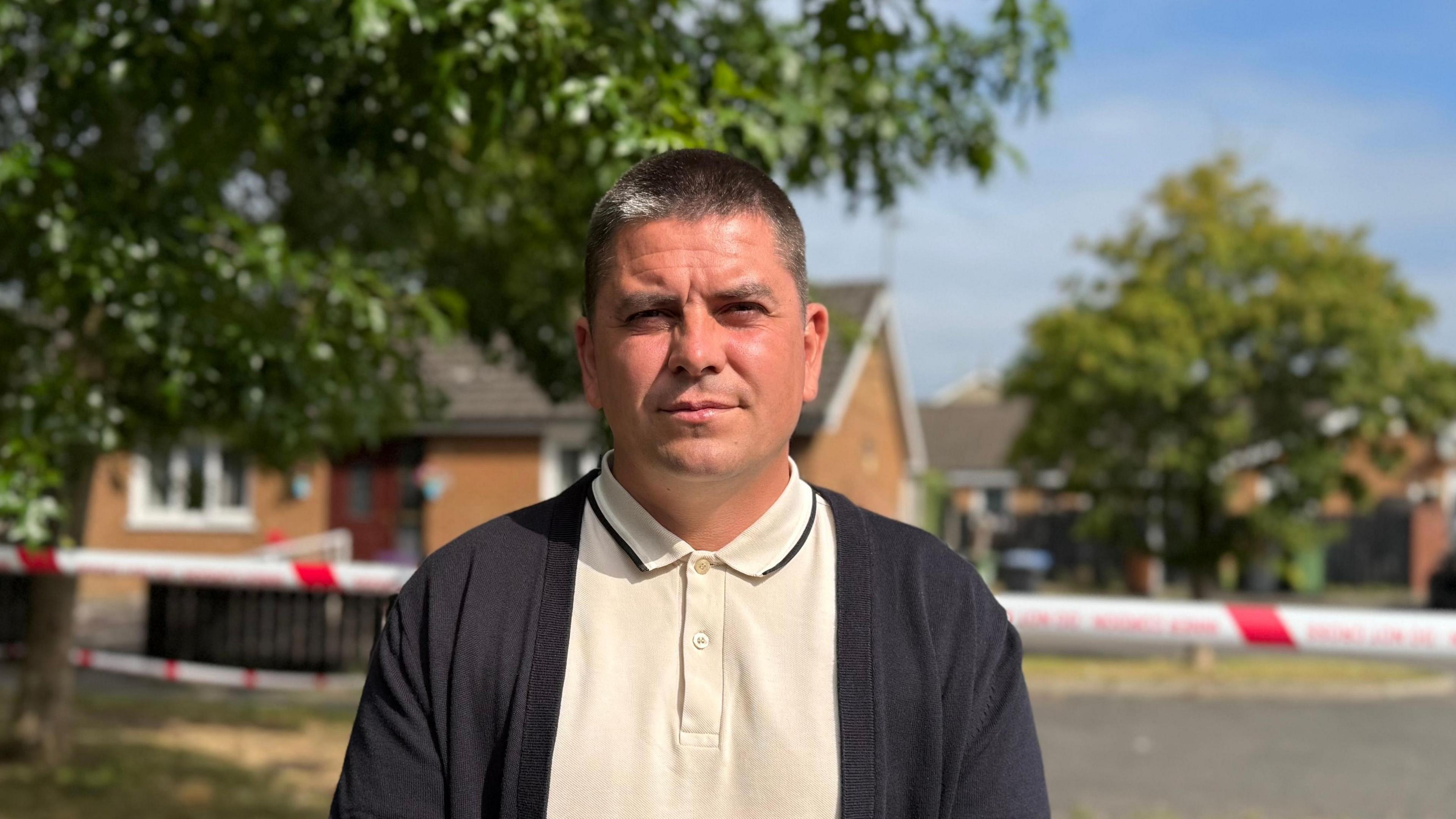 Danny Baker has short black hair with a grey patch. He is staring at the camera as he stands in front of houses where there is red and white tape across the street. He is wearing a beige polo shirt top and a black cardigan.