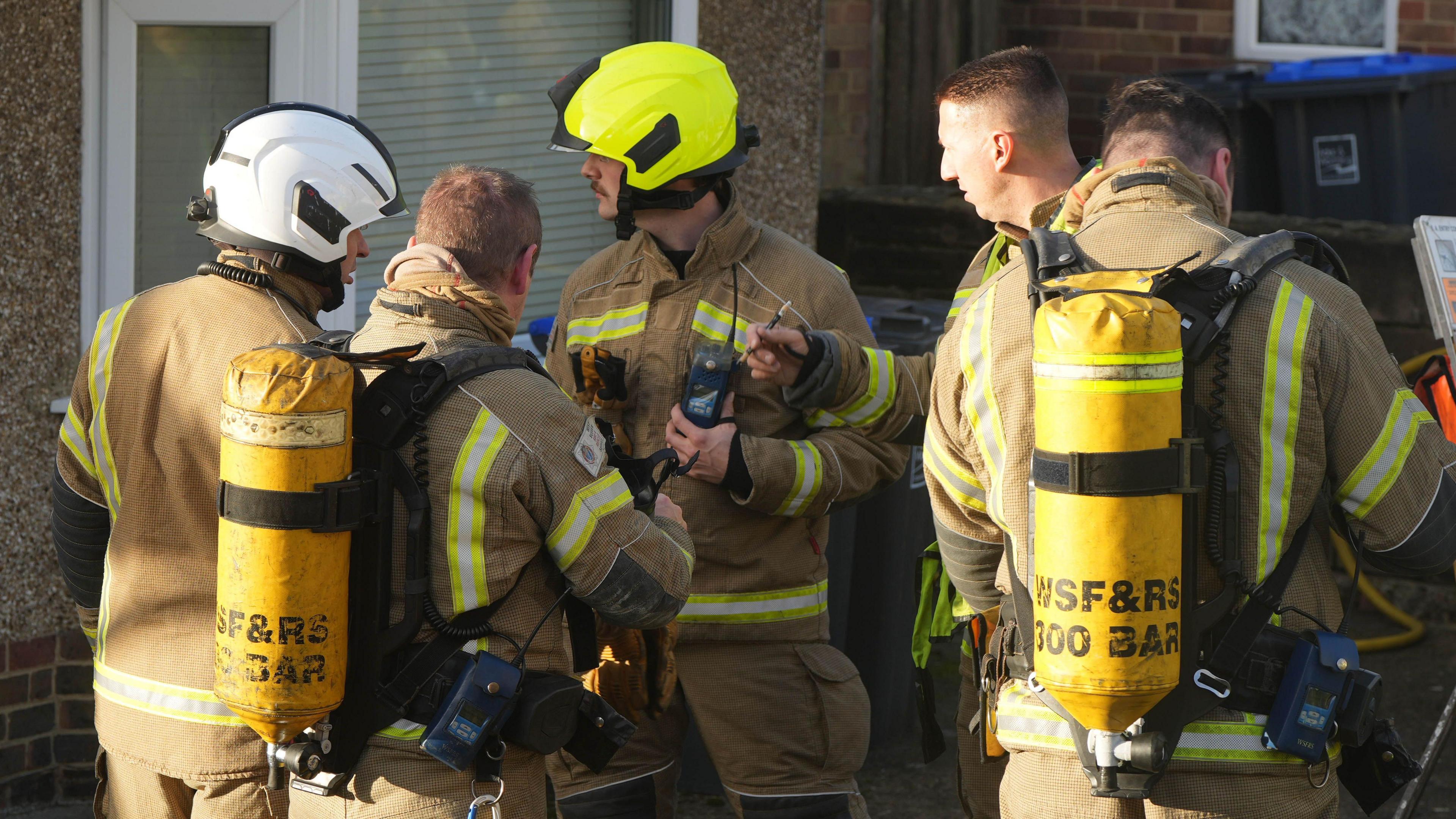 A group of five firefighters stood outside a house. They are all wearing beige uniforms with reflective yellow stripes. Two of them have oxygen cannisters on their backs and two of them are wearing helmets. 