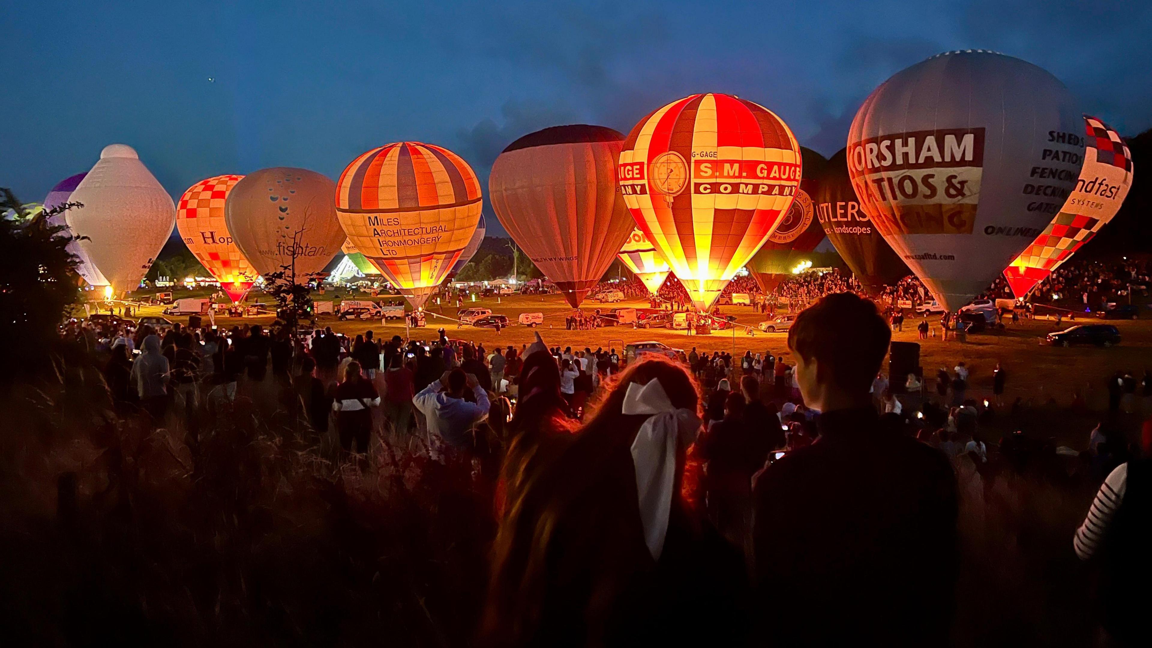 A wide shot of the nightglow at the Bristol Balloon Fiesta showing tethered balloons being illuminated by their burners, with massed crowds visible in the foreground