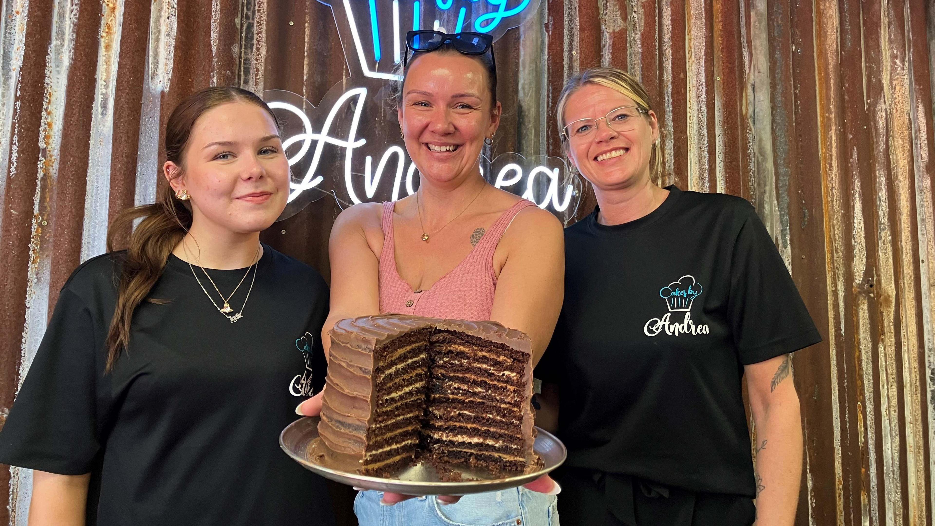 Three women are smiling at the camera. On the left, a woman with long brown hair is wearing a dark T-shirt and two necklaces. A woman wearing a peach vest and sunglasses on her head is in the middle, holding a chocolate cake on a plate. On the right is a woman with long blonde hair and a dark T-shirt with "Andrea" written on it. There is an illuminated cake sign and the word "Andrea" on the wall behind them.
