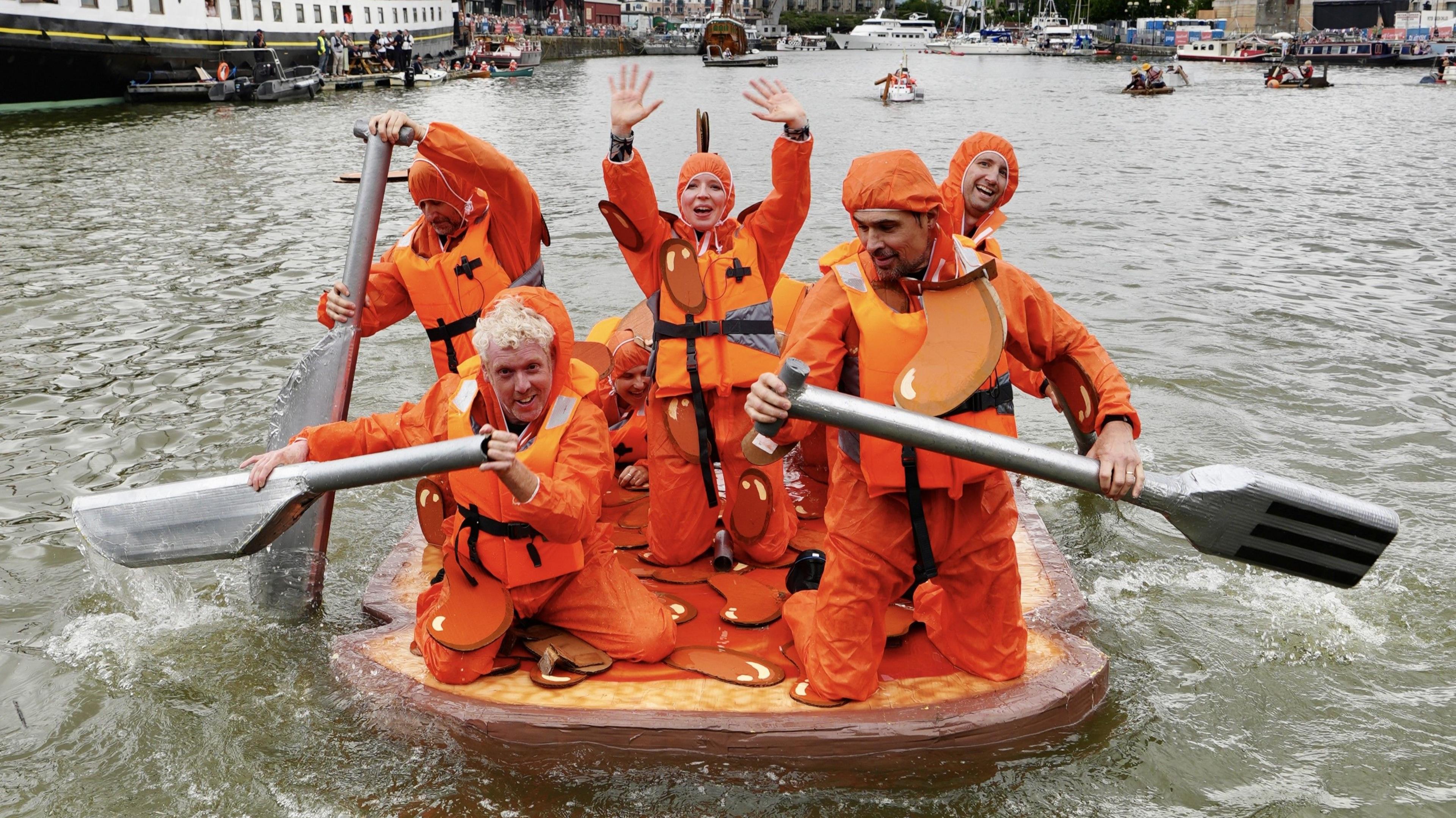 Six people dressed in bright orange overalls, paddling across Bristol's harbourside on a raft, using large oars which have been designed to look like knives and forks. The raft they are floating on looks like a piece of toast, and they are meant to resemble beans.