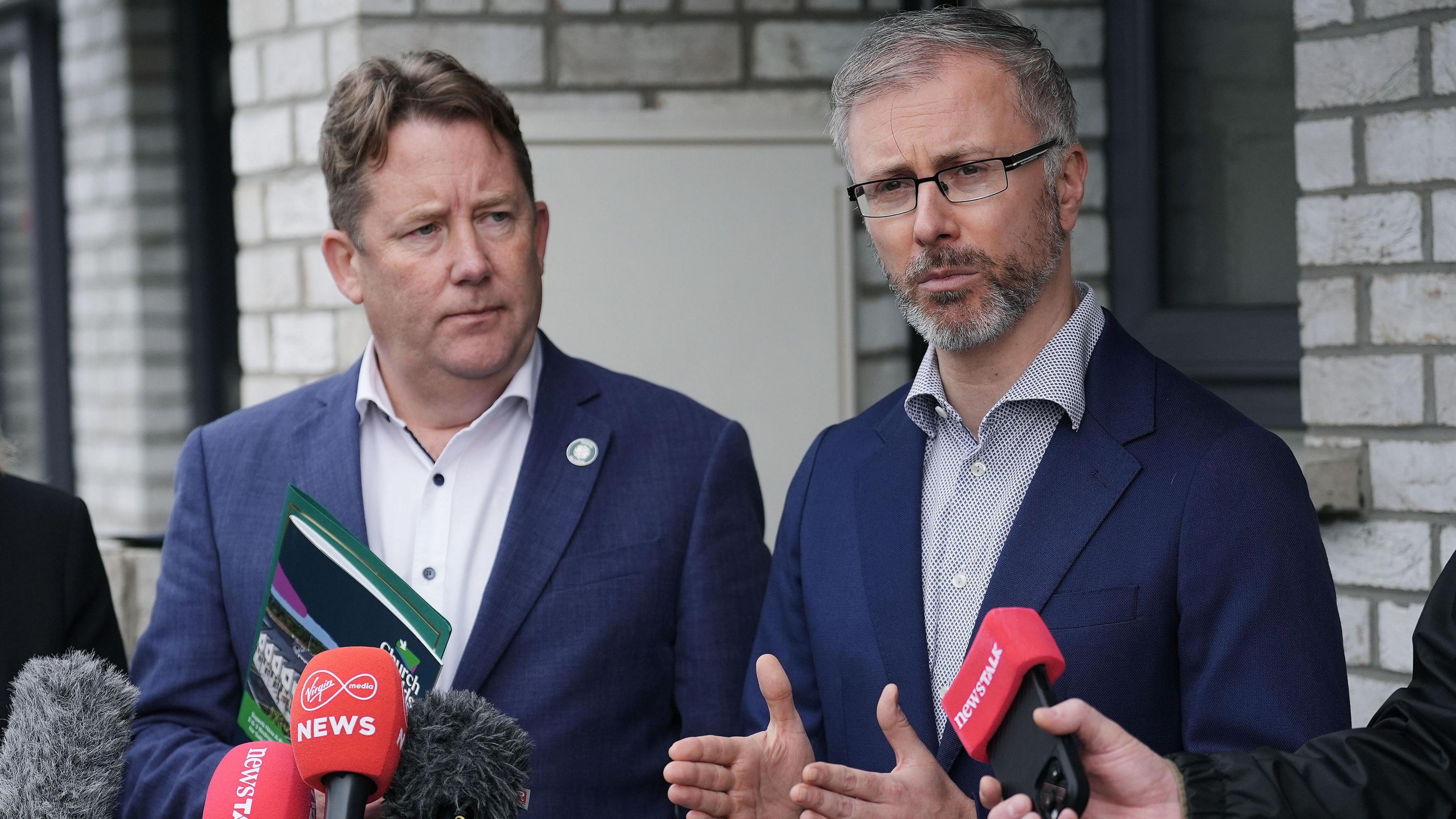 Darragh O'Brien stands to the left of Roderic O’Gorman. O'Brien is looking at O'Gorman while holding a booklet. He is wearing a navy suit jacket and white collared shirt with the top button undone. On the right O'Gorman is speaking into microphones. He has grey hair and beard, is wearing rectangle frame black glasses and wearing a navy suit jacket and patterned collared shirt.