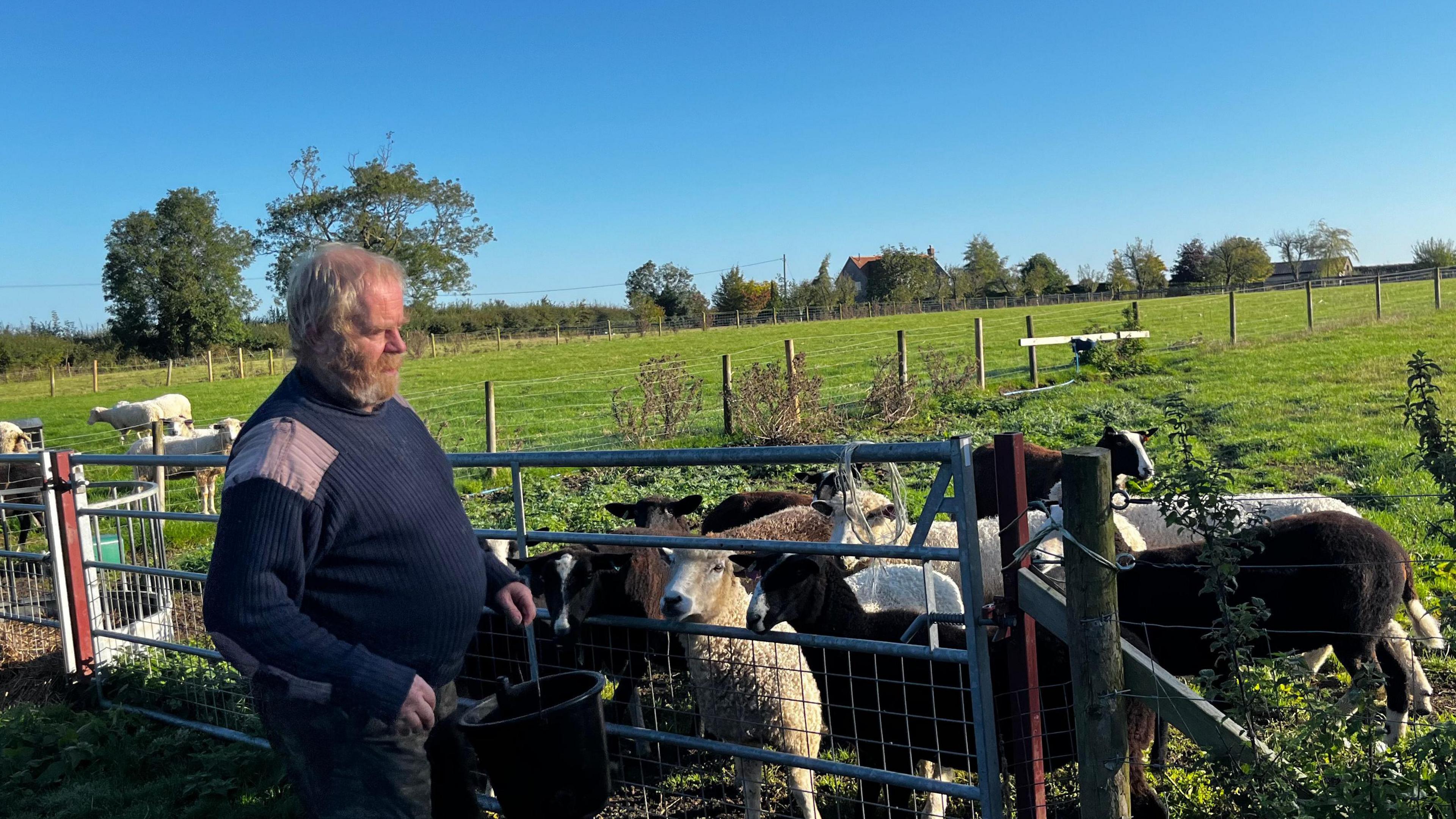 A man in a woolly jumper and combat trousers stands in front of a field of sheep.