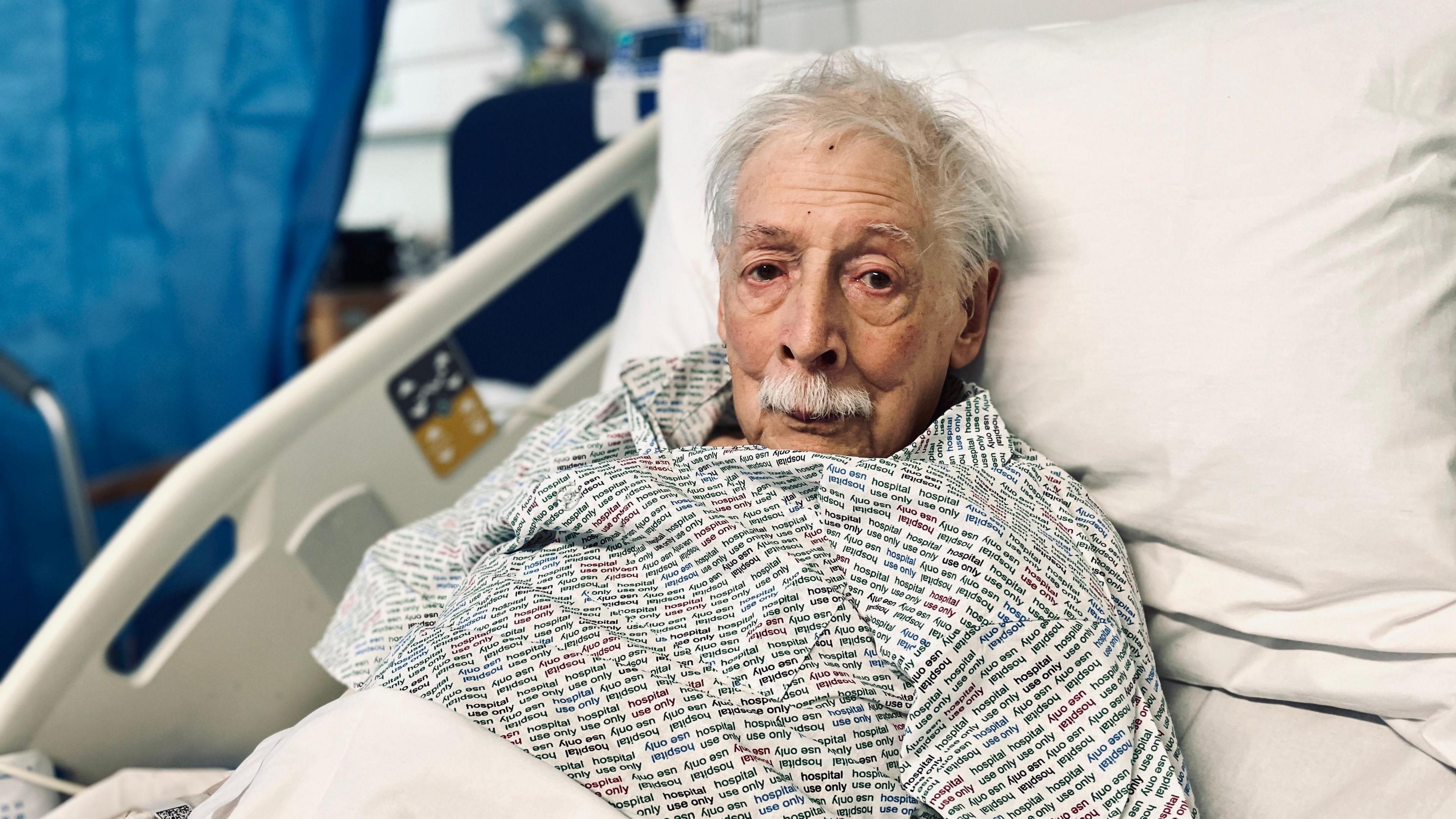 Michael Foster looks at the camera. He has grey hair and a grey moustache and is in a hospital bed propped up by pillows.