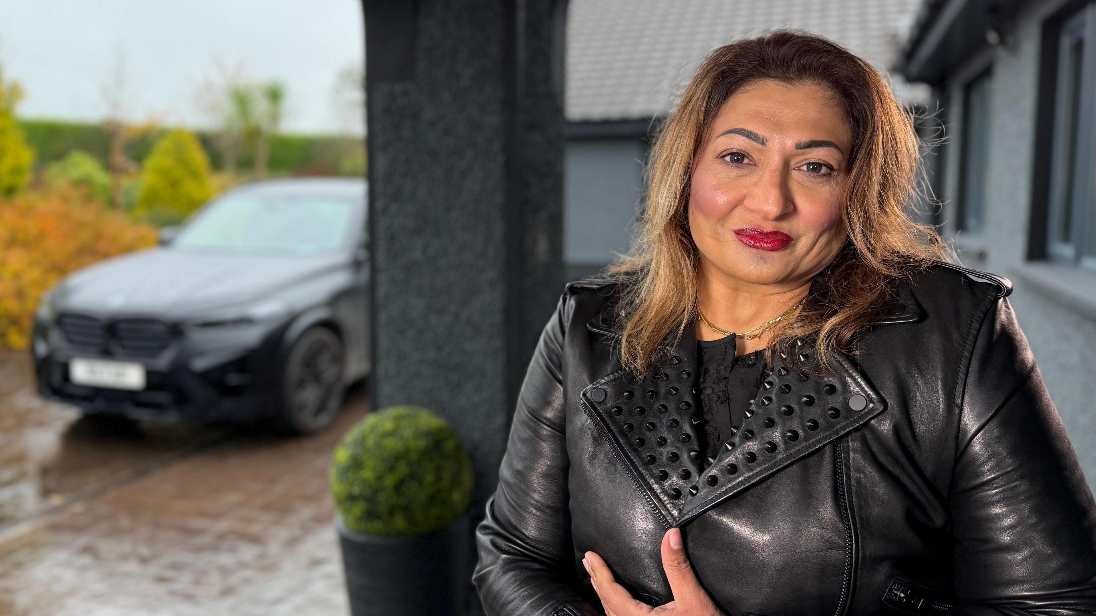 A woman in leather jacket sits in a wheelchair looking into the camera on her front porch. In the background a black jeep is parked in the driveway.