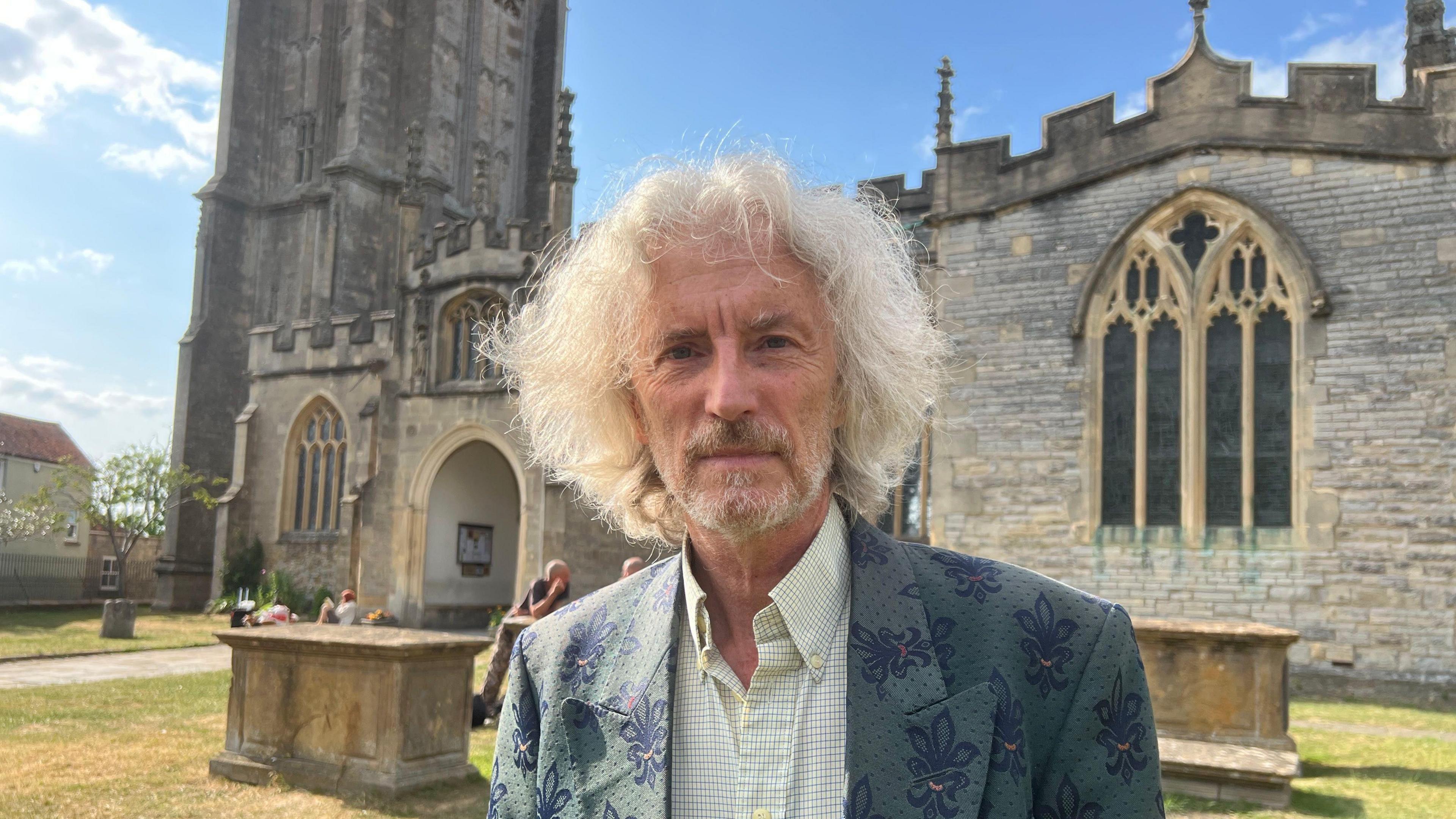 Man in a green suit and shirt with long hair. He's stood in a churchyard with a historic church behind.