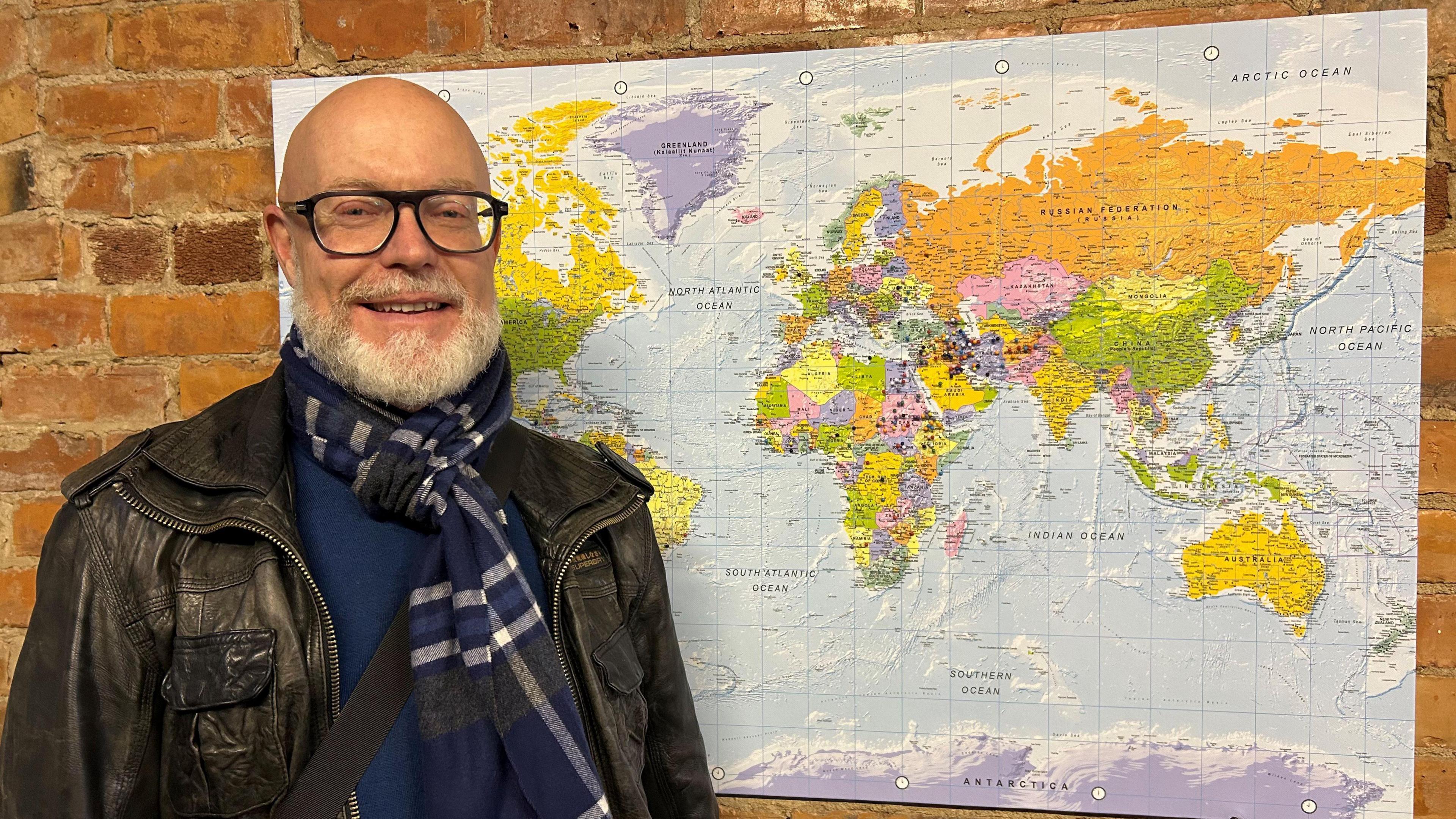 A bald man with glasses and wearing a blue scarf and leather jacket, stands next to a map of the world, which has been placed on a wall. Small pins have been placed on some of the countries on the map.