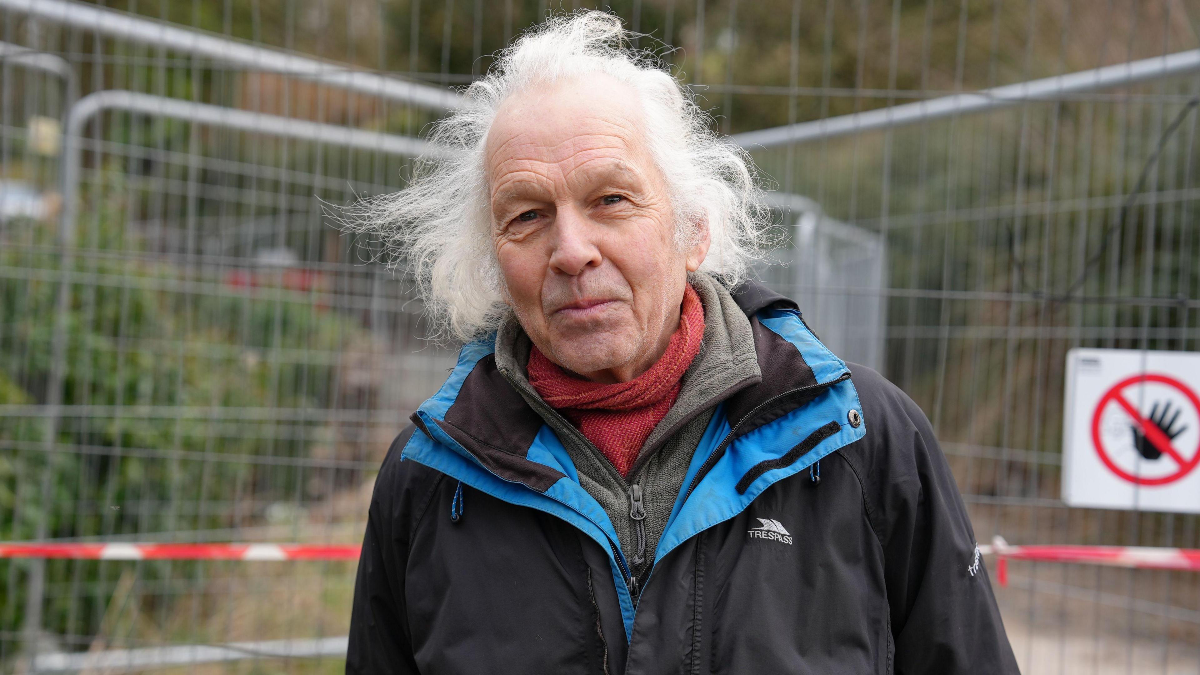 Simon Greaves, who has long white hair, stands in front of a metal barrier which is closing the road in Bronygarth. He is wearing a black coat with blue collar, a red scarf and green fleece. 