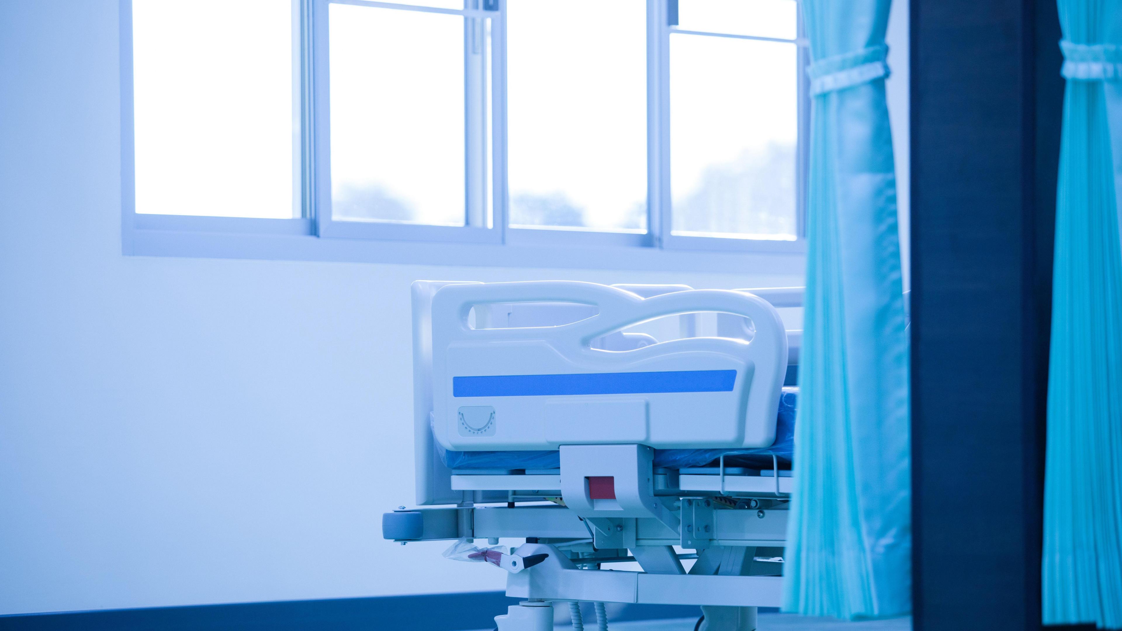 The bottom of an empty hospital bed. A blue curtain is pulled closed. A window is next to the bed.