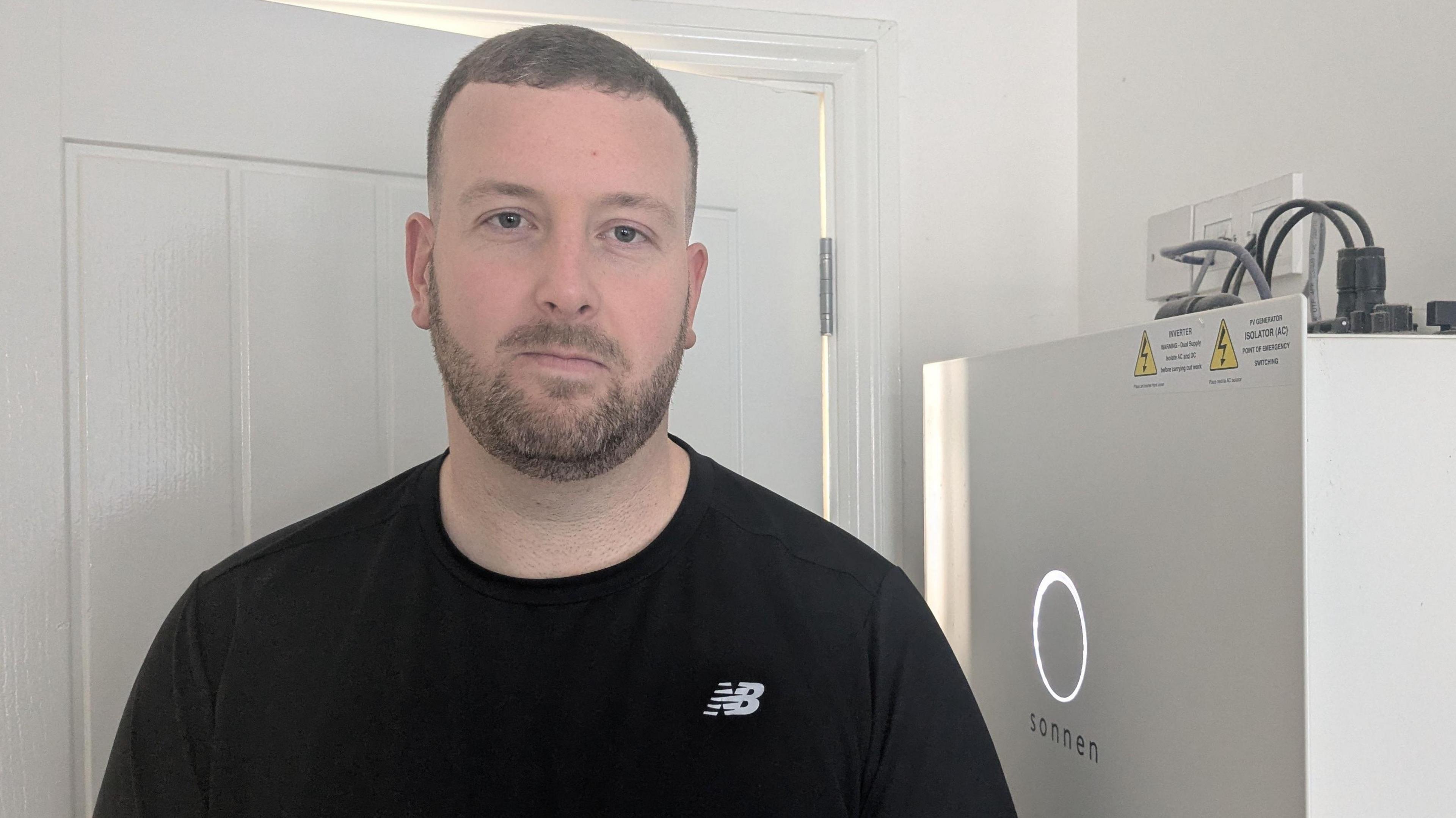 A man with a beard stands in front of his home heating system. He's wearing a black t shirt and has short brown hair. Next to him is a white appliance with a white ring of light on the front.