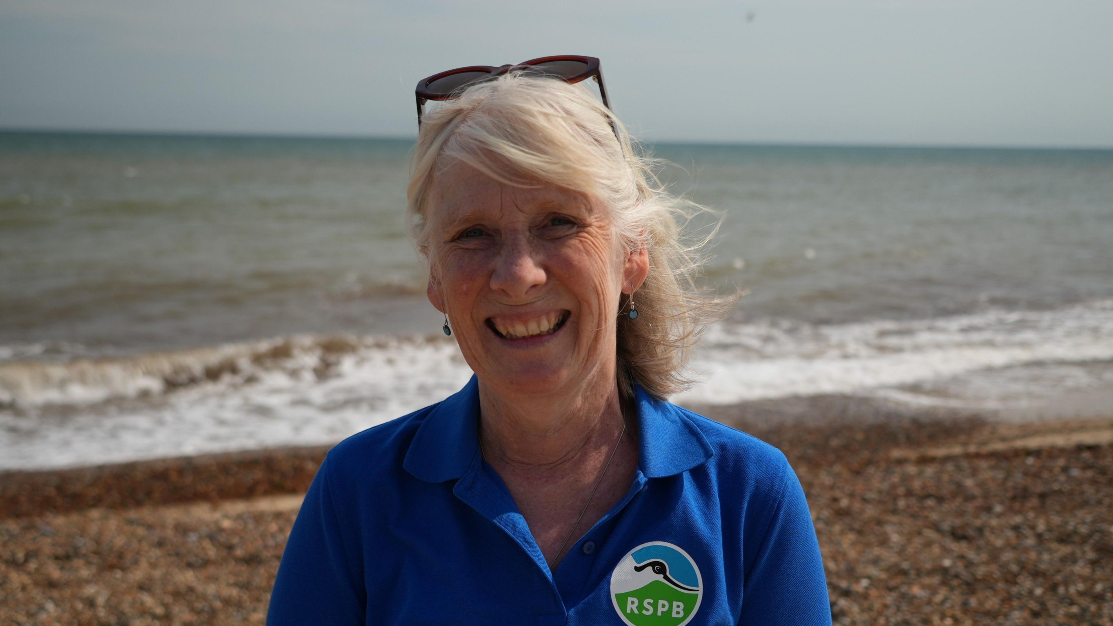 A woman smiles at the camera in a head and shoulders image. She is wearing a blue polo shirt with a round RSPB badge to the left of her chest, featuring a black and white avocet. The woman has grey hair and sunglasses perched on top of her head.