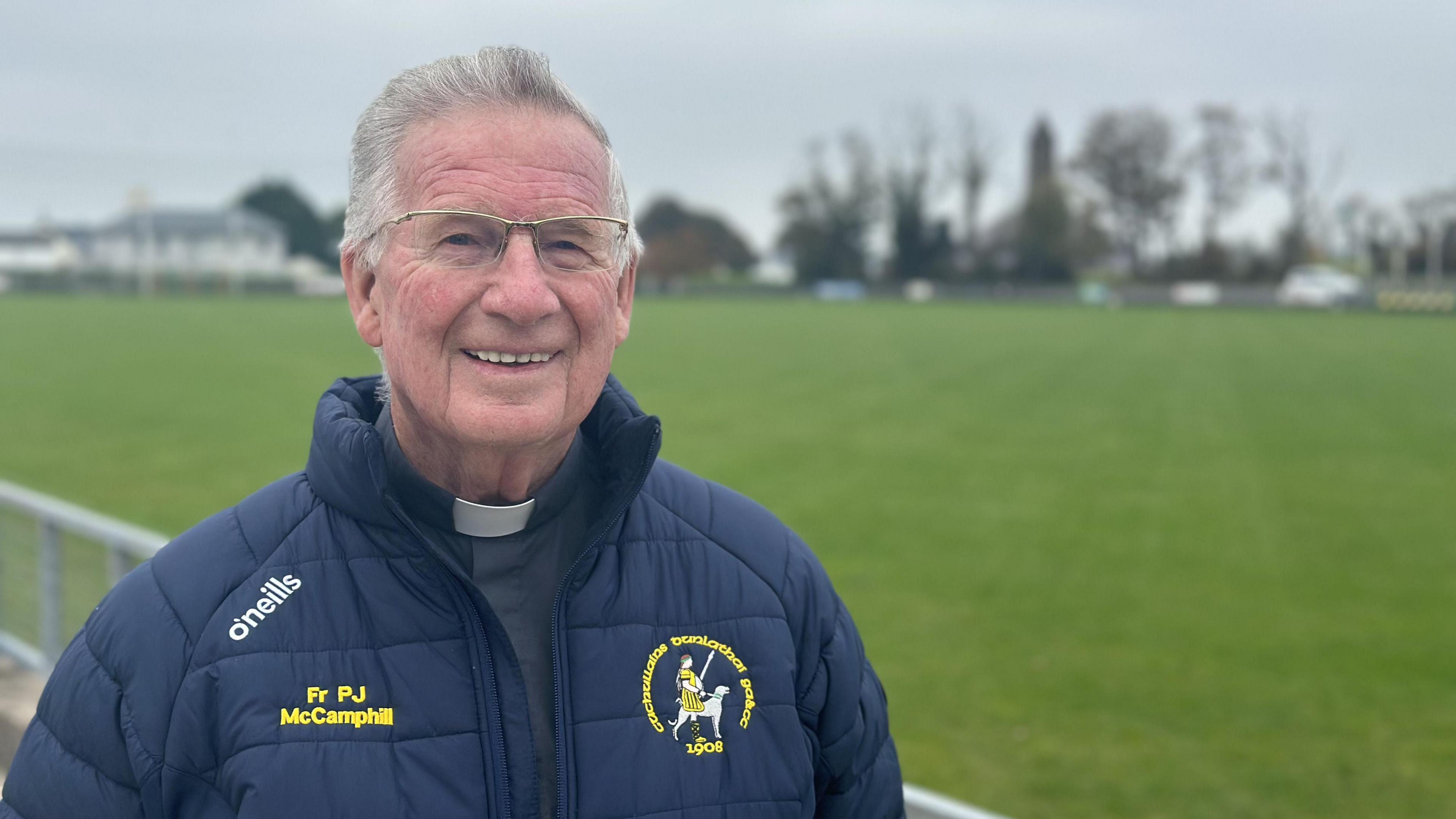 A older man, with grey hair, smiling at the camera. He is wearing a dark grey top, a white clerical collar and a navy GAA jacket. He is smiling at the camera and is standing in front of a GAA pitch.