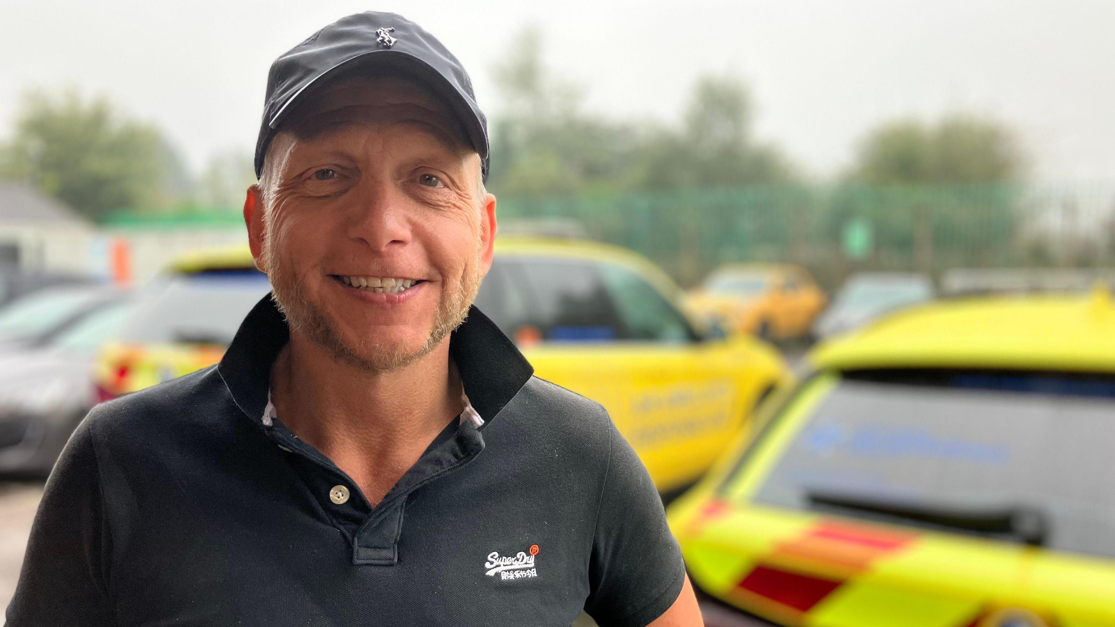 Mike Bray in a black polo shirt and black cap standing in front of emergency vehicles