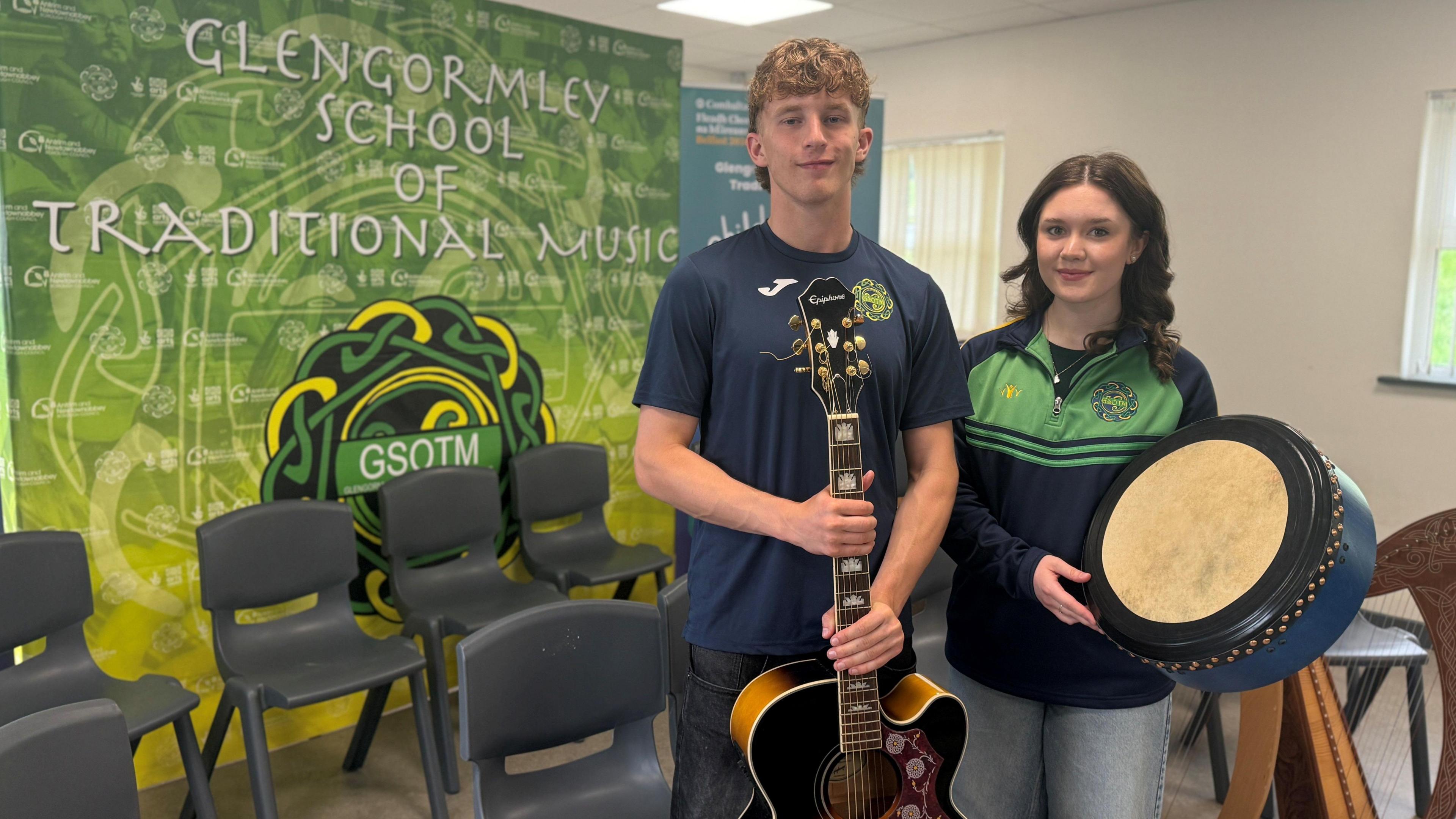 Two teenagers, a male and female, both with long brown hair. Jacob wears a navy uniform tshirt and is holding a guitar. Niamh is also in uniform, a green and navy half zip, holding a bordhan. They are pictured beside grey chairs and a green Glengormley School of Traditional Music backdrop.