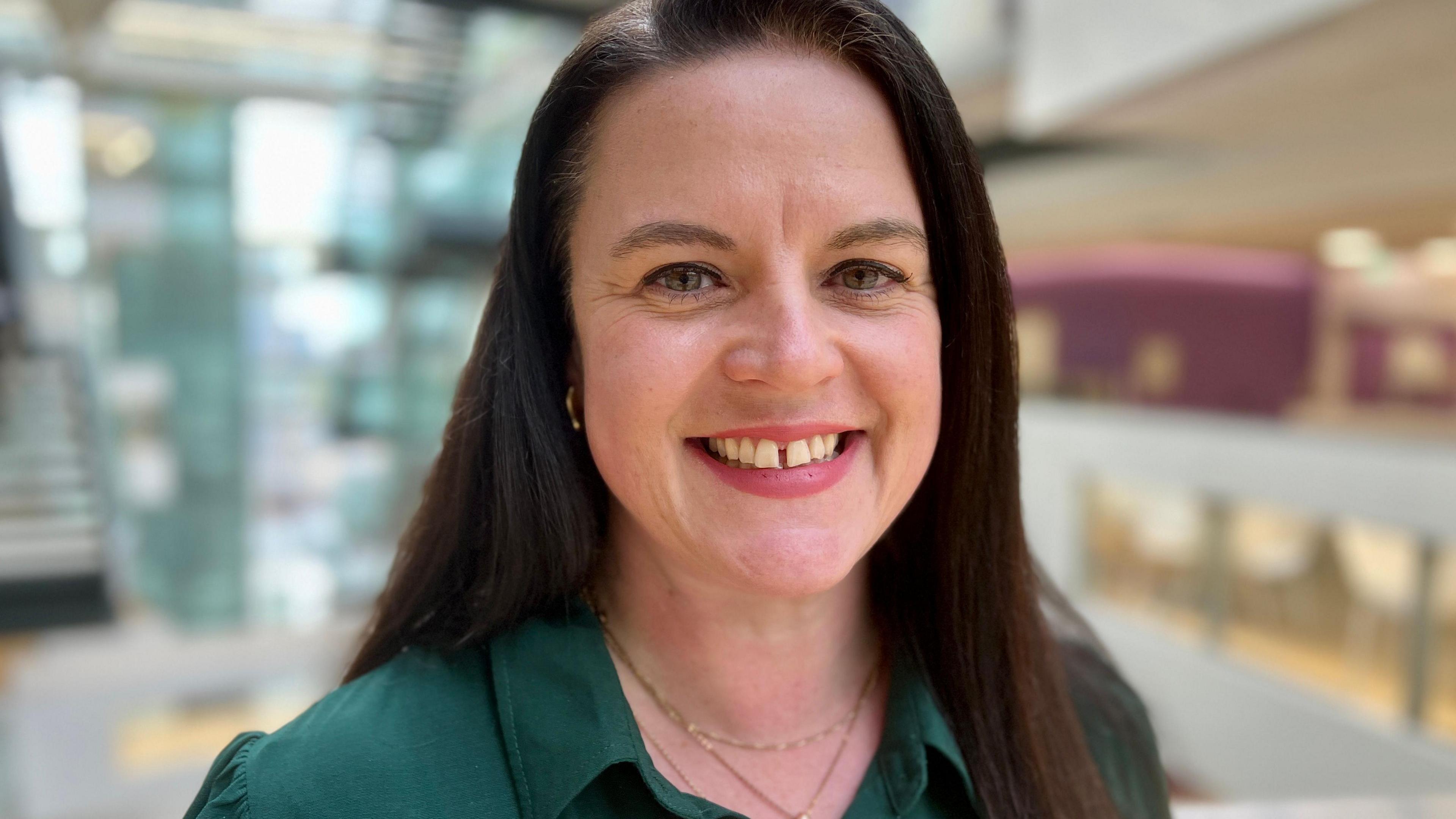 Councillor Beccy Hopfensperger smiles at the camera. She has long dark brown hair and a gap in her front teeth. She is wearing a dark green dress with a collar.