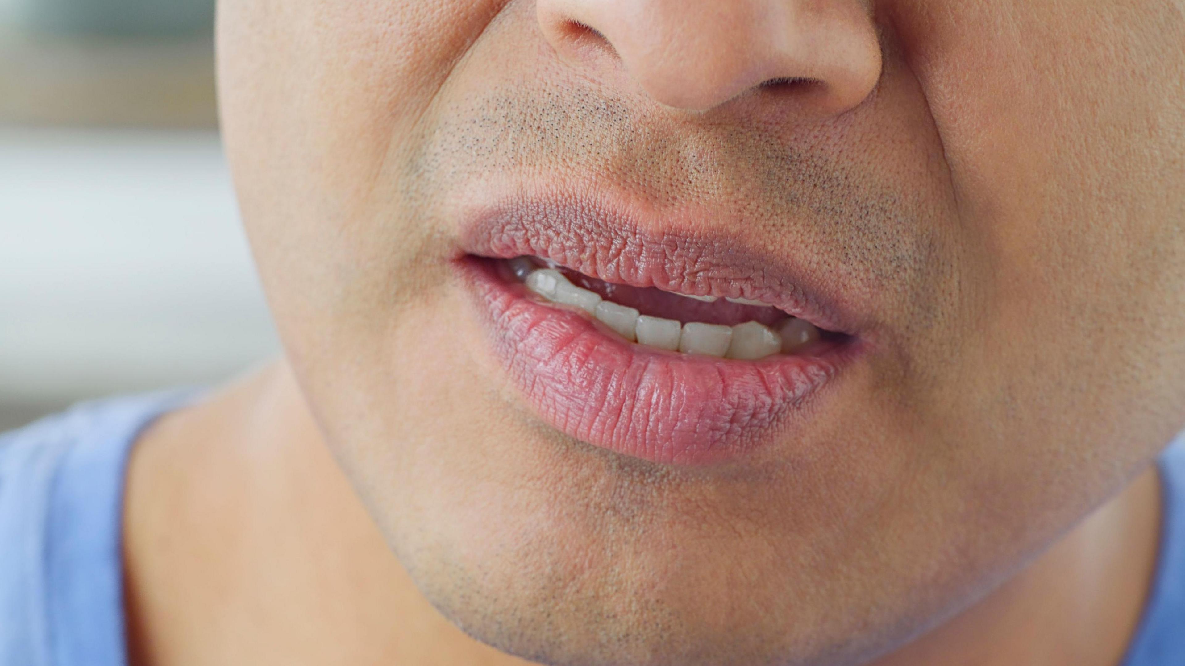 A close-up of a man's mouth and jaw. His lips are partly open and you can see his lower teeth. He is wearing a blue T-shirt