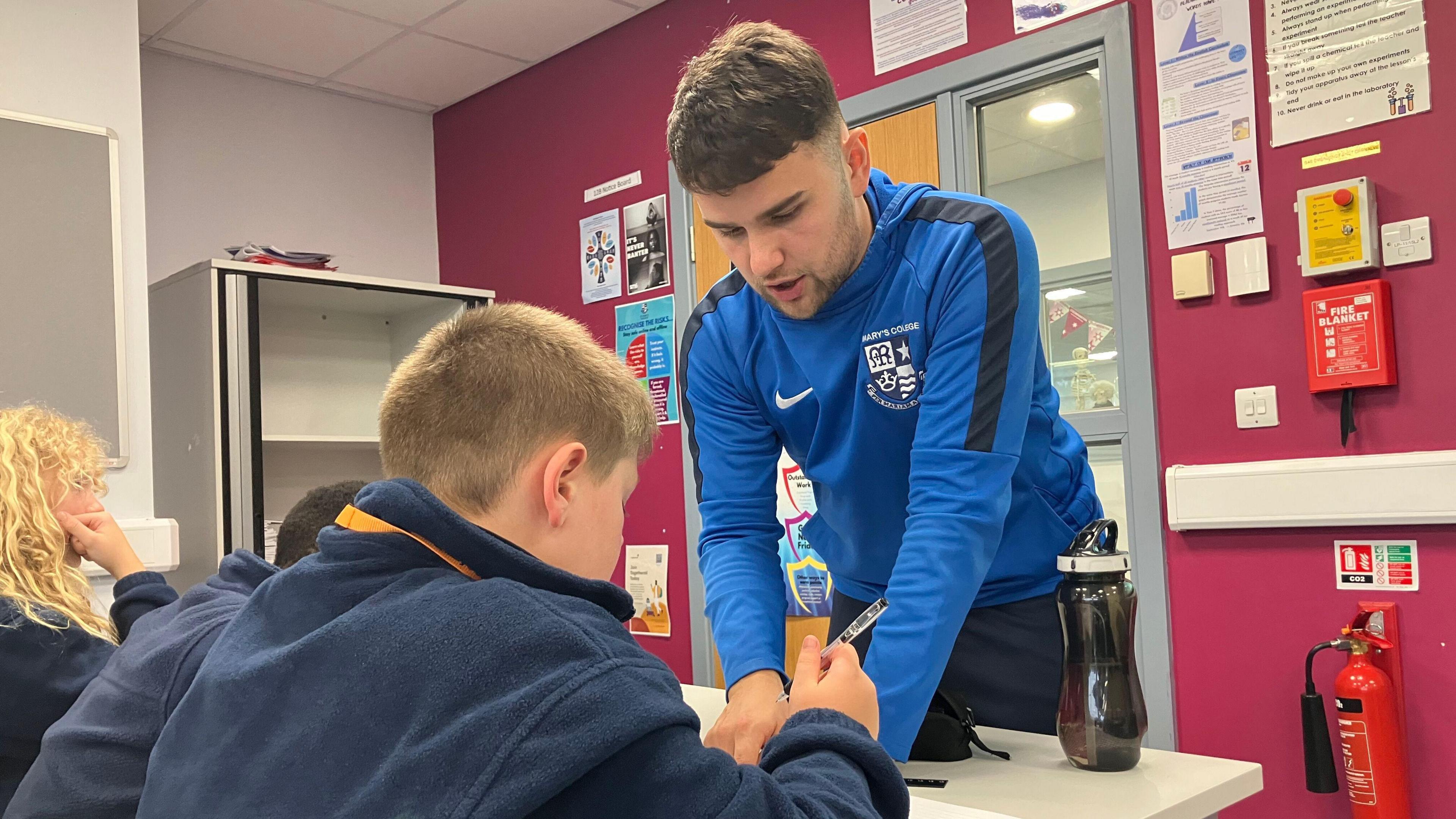 Callum Vernon is in a blue and black tracksuit standing at a student's desk pointing at his work. The student with blonde short hair and a pen in his hands looks at the paper.