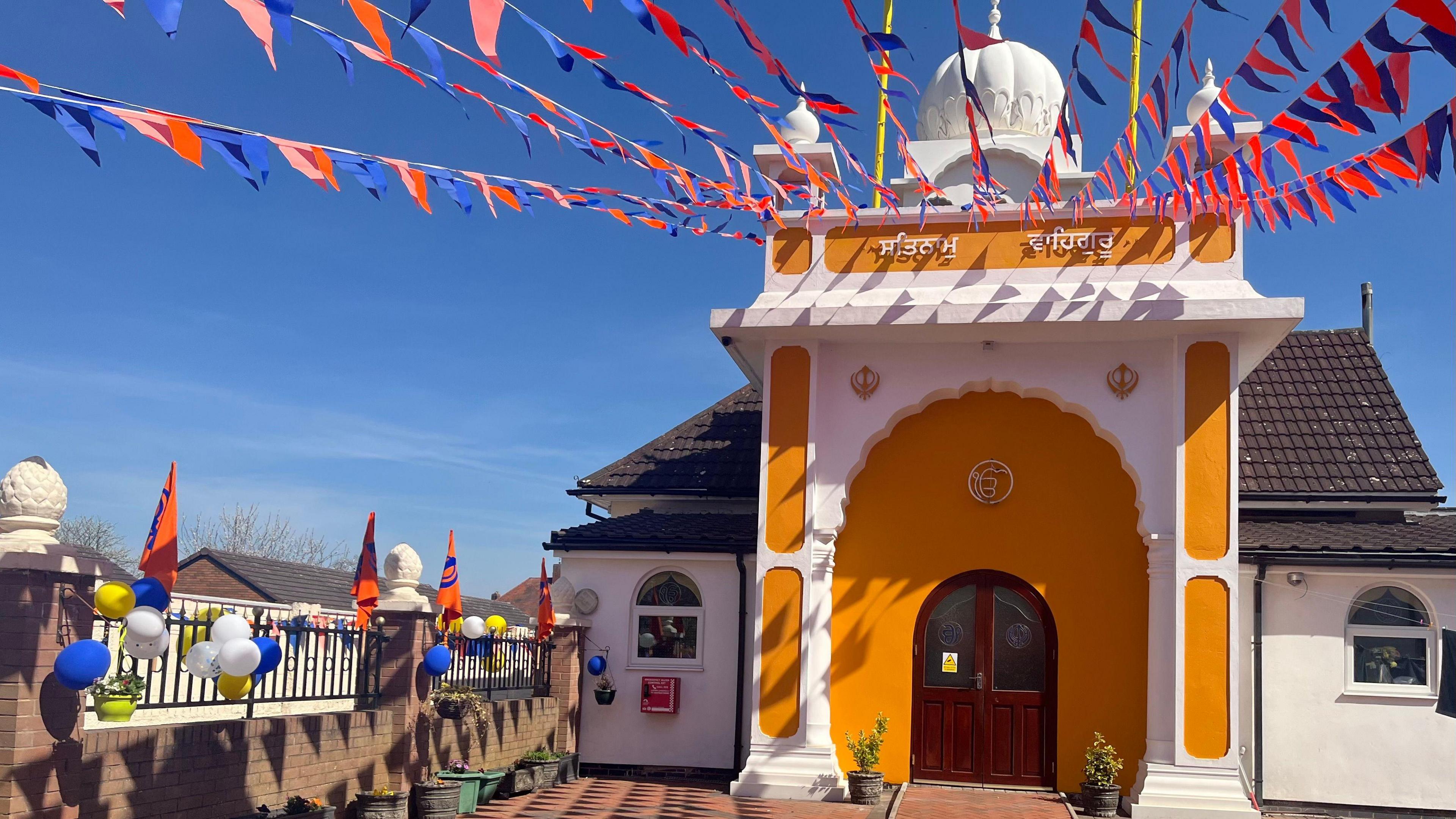The gurdwara is painted orange and white. There are flags hanging from the ceiling of the gurdwara and a blue sky can be seen in the background.