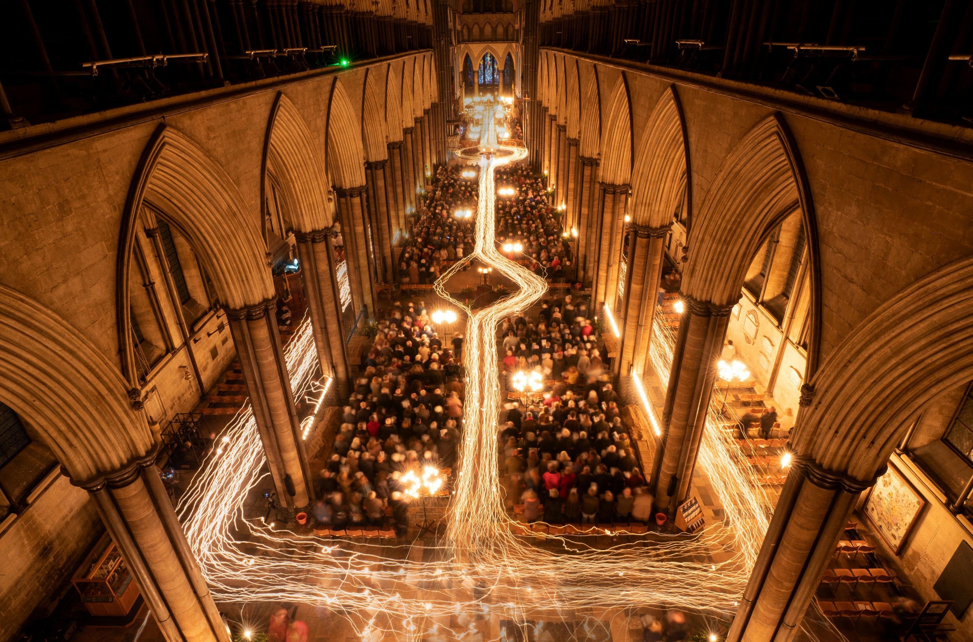 An aerial view of the inside of Salisbury Cathedral during the Advent darkness to light service. Hundreds of people and the choir can be seen holding candles while a timelapse effect shows the path the choristors took at the start of the service