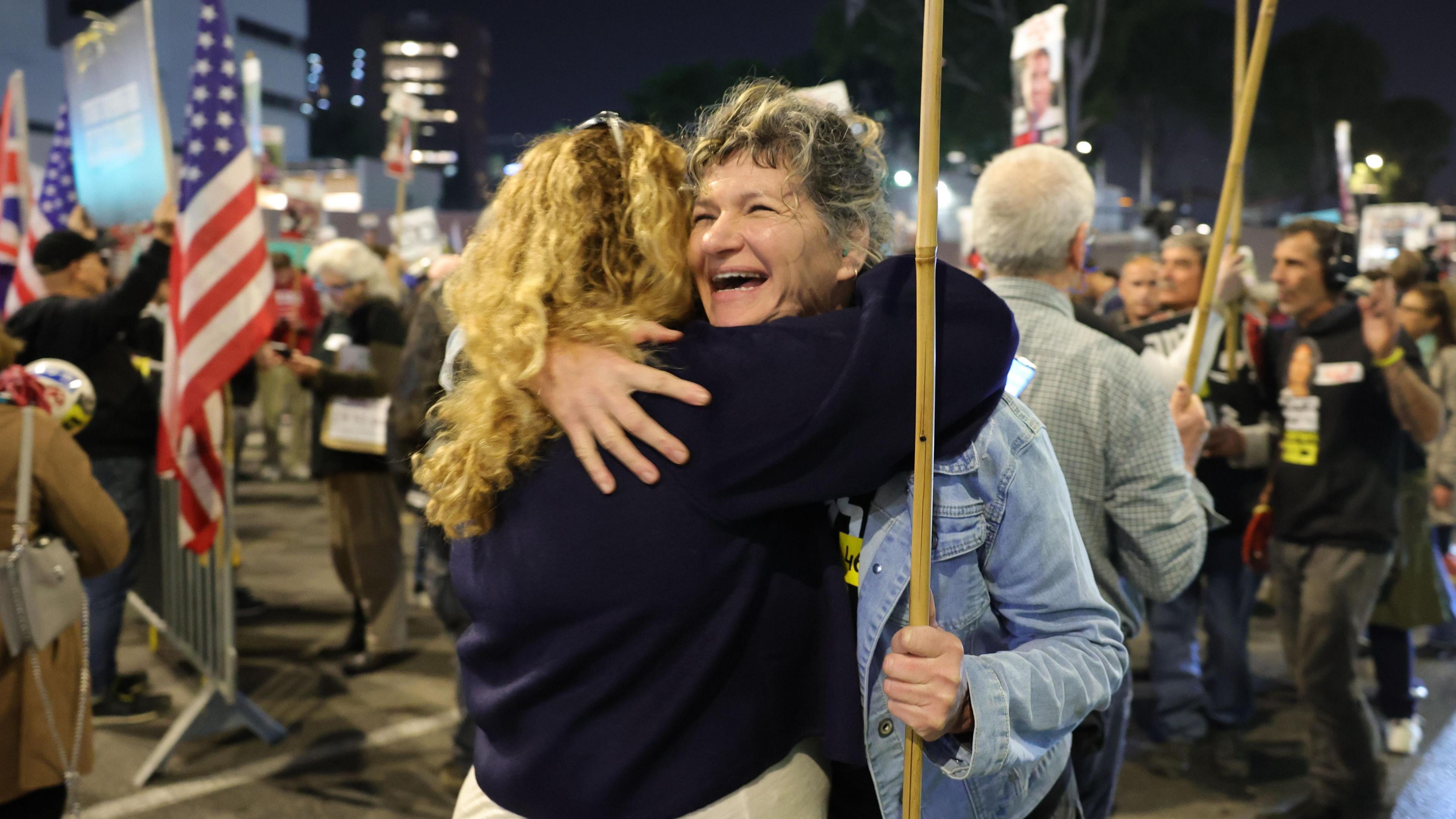 Two women hug in the middle of crowded protest at night. The woman facing the camera is smiling while holding a placard.