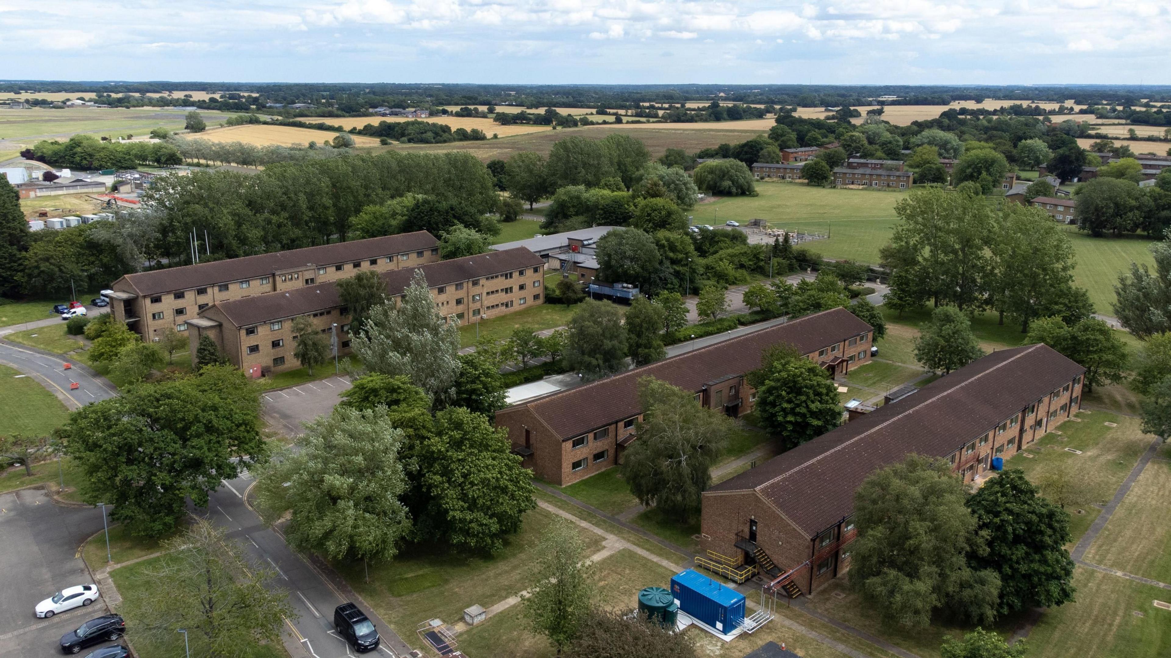 Aerial shot of the housing blocks at an airbase. Four rectangular accommodation blocks can be seen, surrounded by grass, trees and roads. There are agricultural fields in the background.