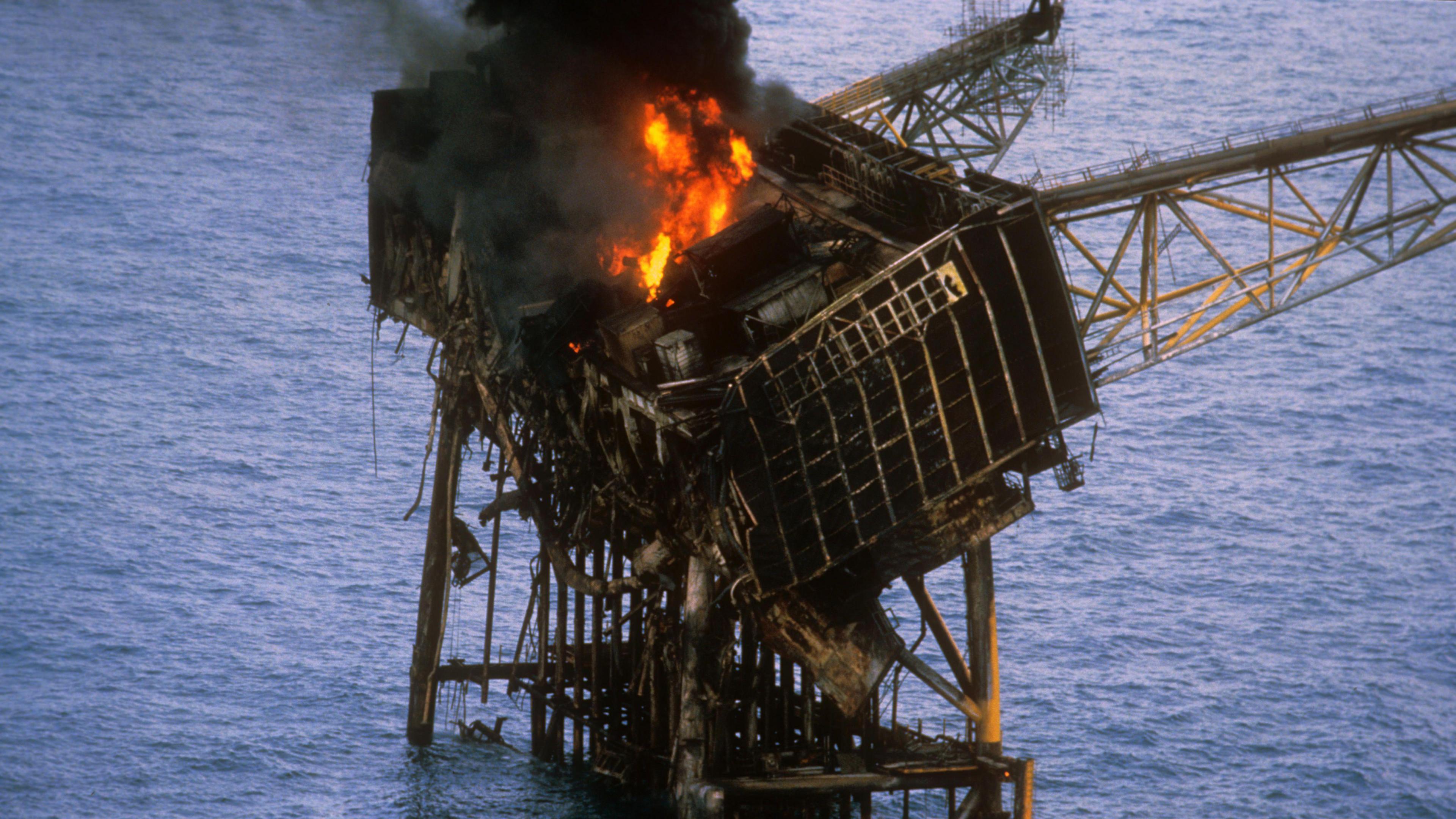 The badly-buckled and blackened platform is still aflame in the daylight the day after the disaster. The smoke coming from the flames is very thick and black. The sea is calm and blue.