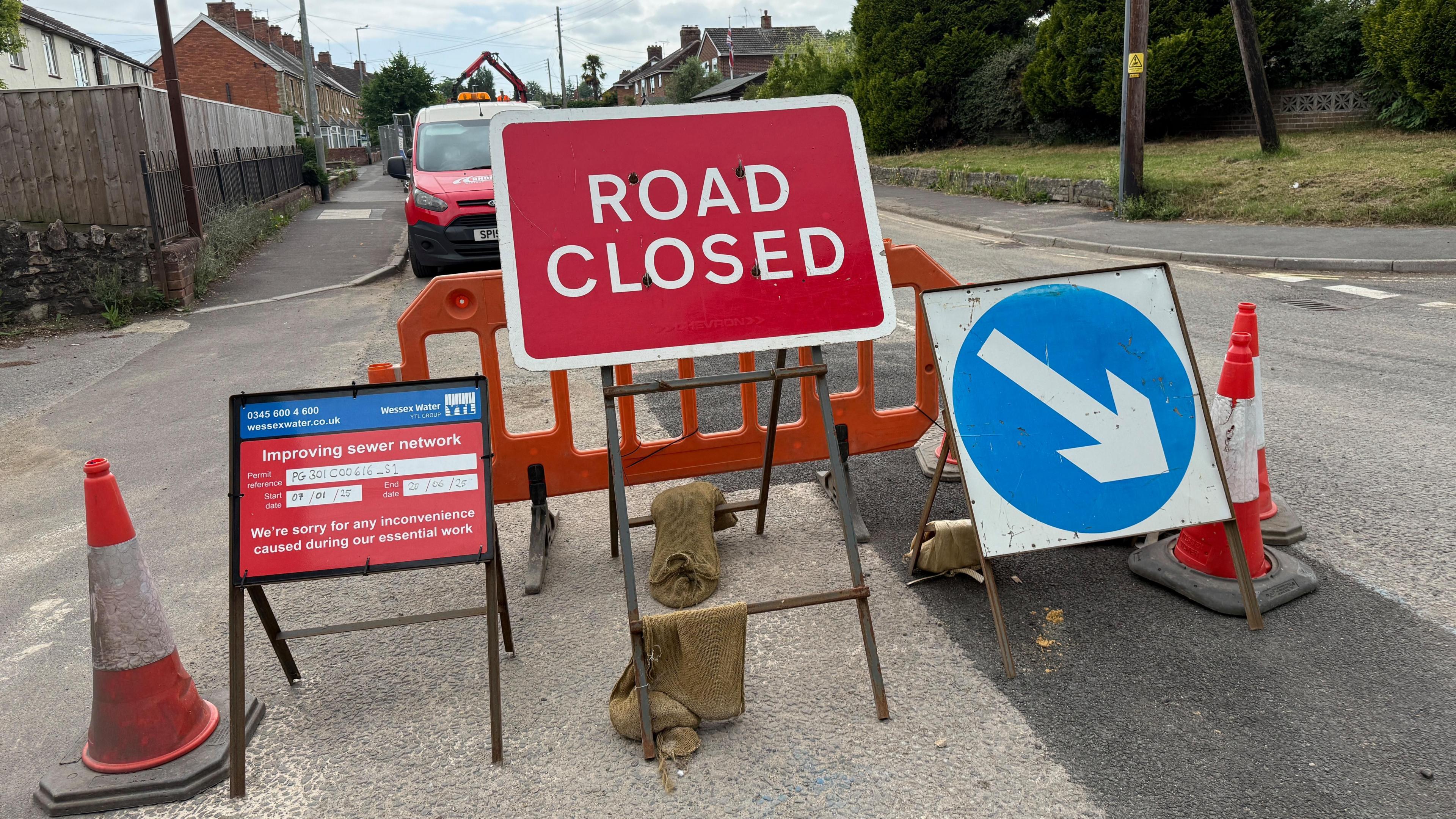 a red road closed sign with other signs and traffic cones in the middle of a road with a strip of fresh tarmac