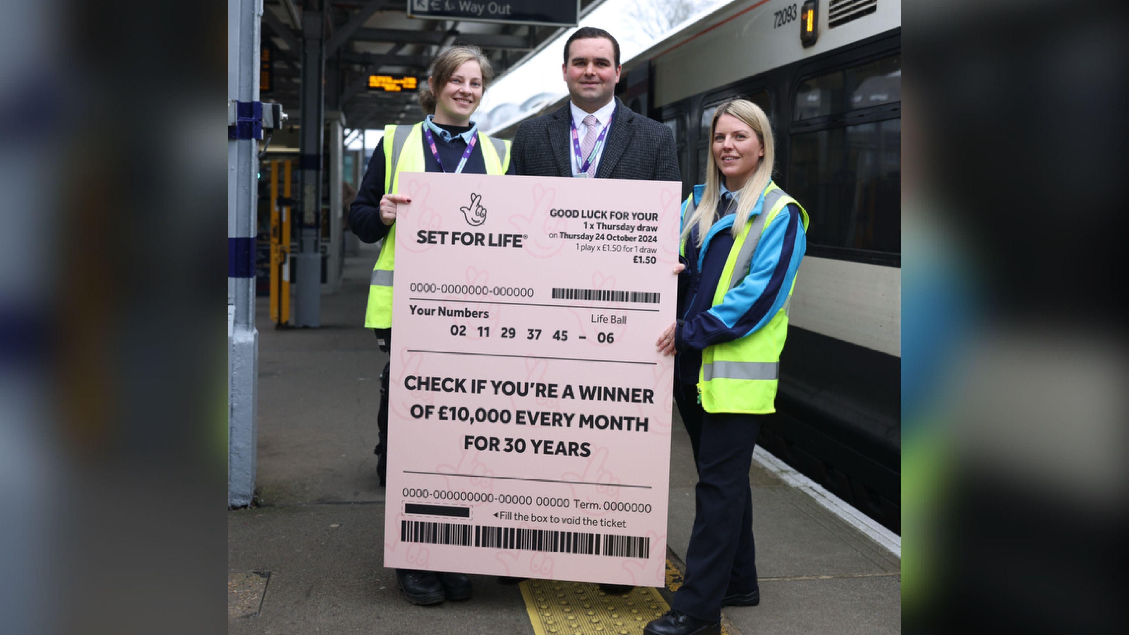 Two women in yellow hi-vis jackets and Southeastern uniforms standing either side of a man in a grey suit. They are holding a giant lottery ticket which says "CHECK IF YOU'RE A WINNER OF £10,000 EVERY MONTH FOR 30 YEARS".