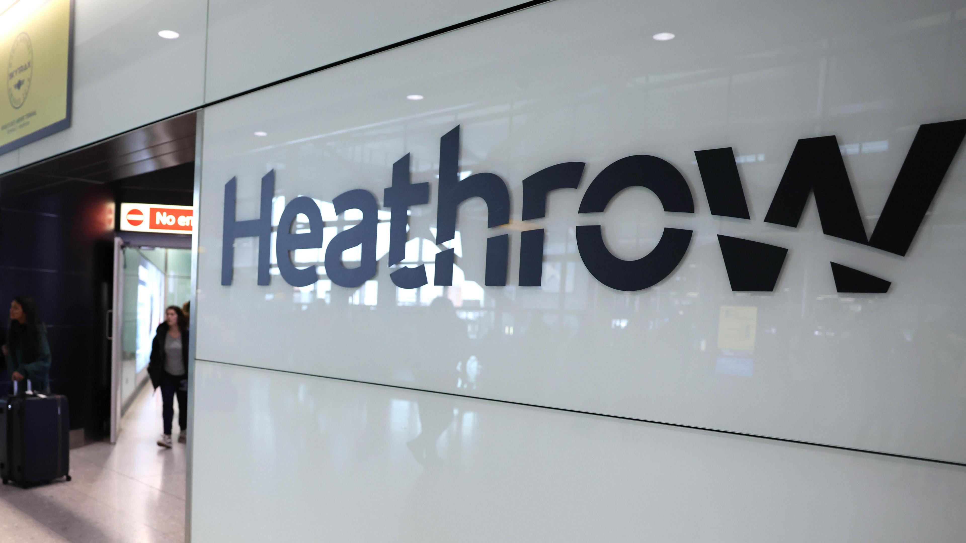 Sign reading Heathrow inside airport terminal. There is an unidentified woman, who is out of focus, walking towards the sign in the background.
