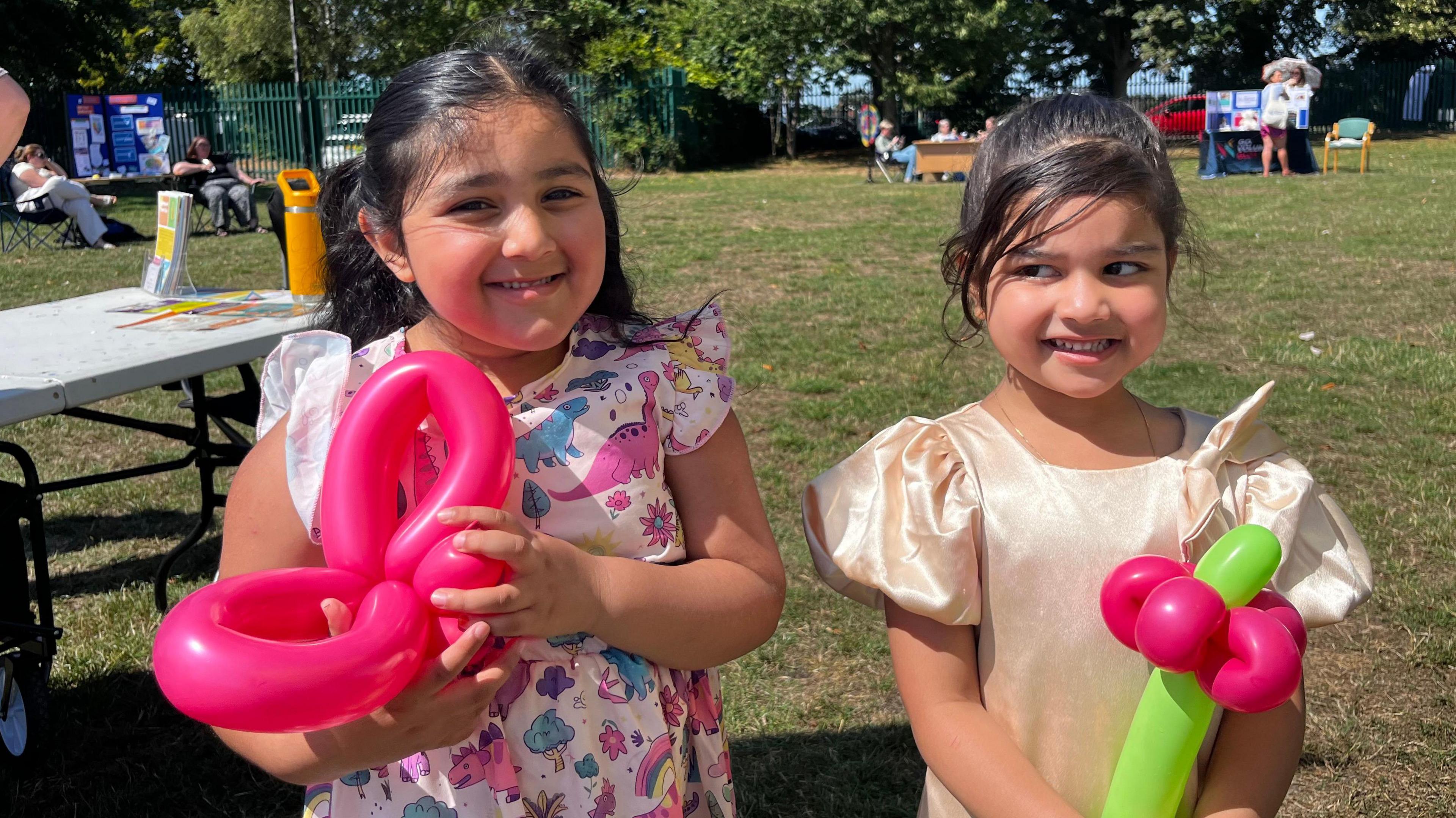 The image features two young girls holding balloon toys. The girl on the left is dressed in a vibrant dinosaur-print dress and is holding a pink butterfly-shaped balloon. Beside her, the girl on the right wears a gold dress and carries a balloon shaped like a flower. They are standing in a grassy field, with a table visible in the background to the left.