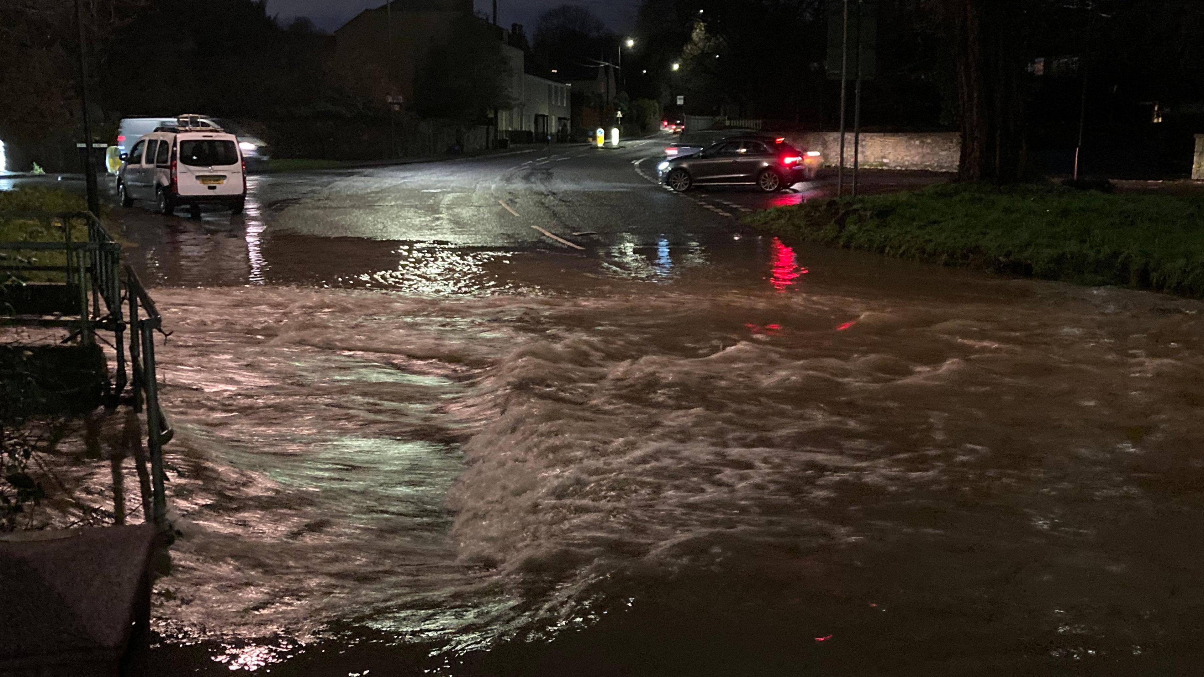 A fast flowing ford in Bristol is flooded with water. The photo is taken in the dark and water can been seen across the road. There are houses and three cars in the background. 