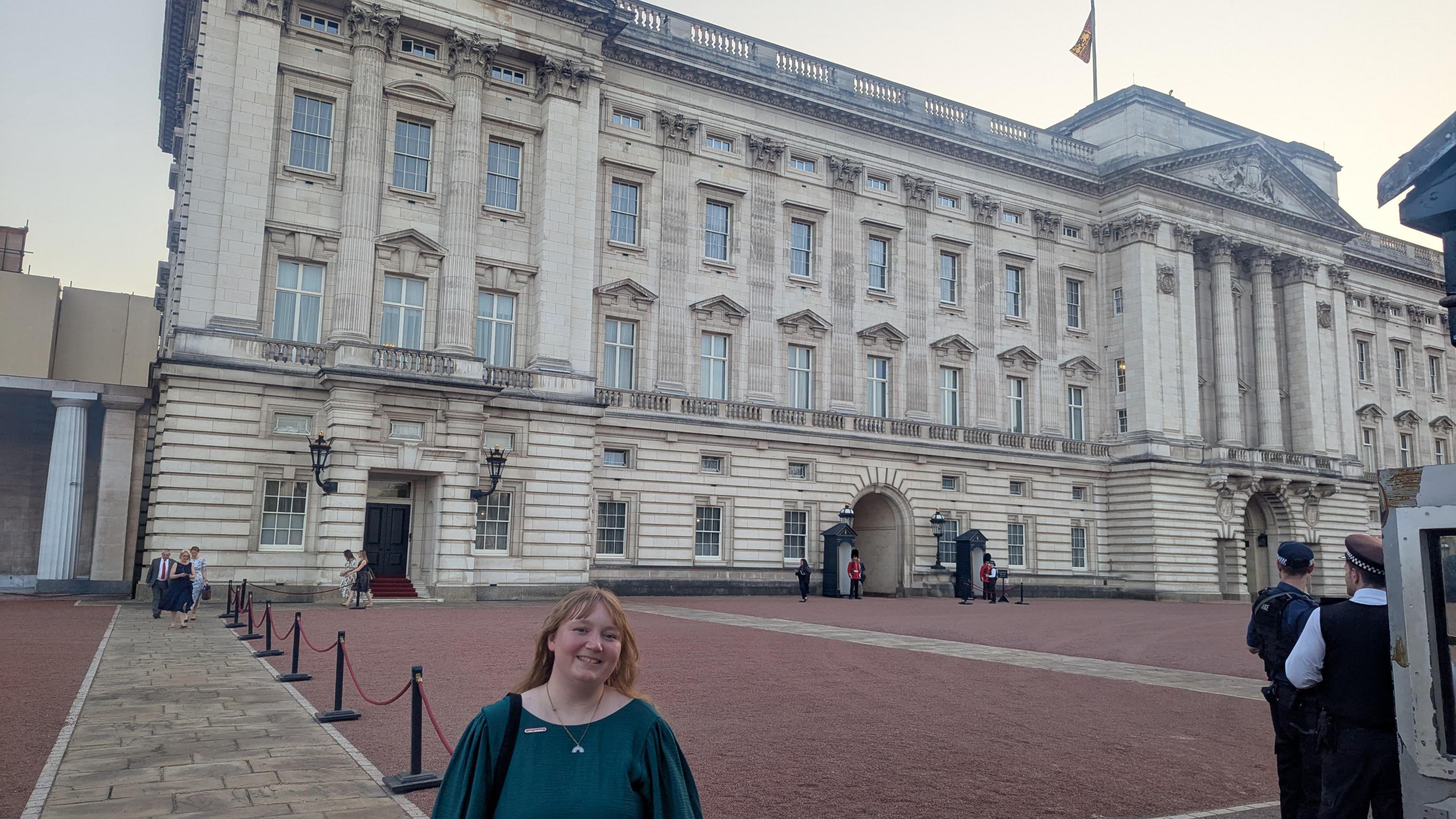 A wider view of Belinda Bradley standing outside Buckingham Palace. The building has multiple storeys with dozens of windows. She is smiling at the camera.