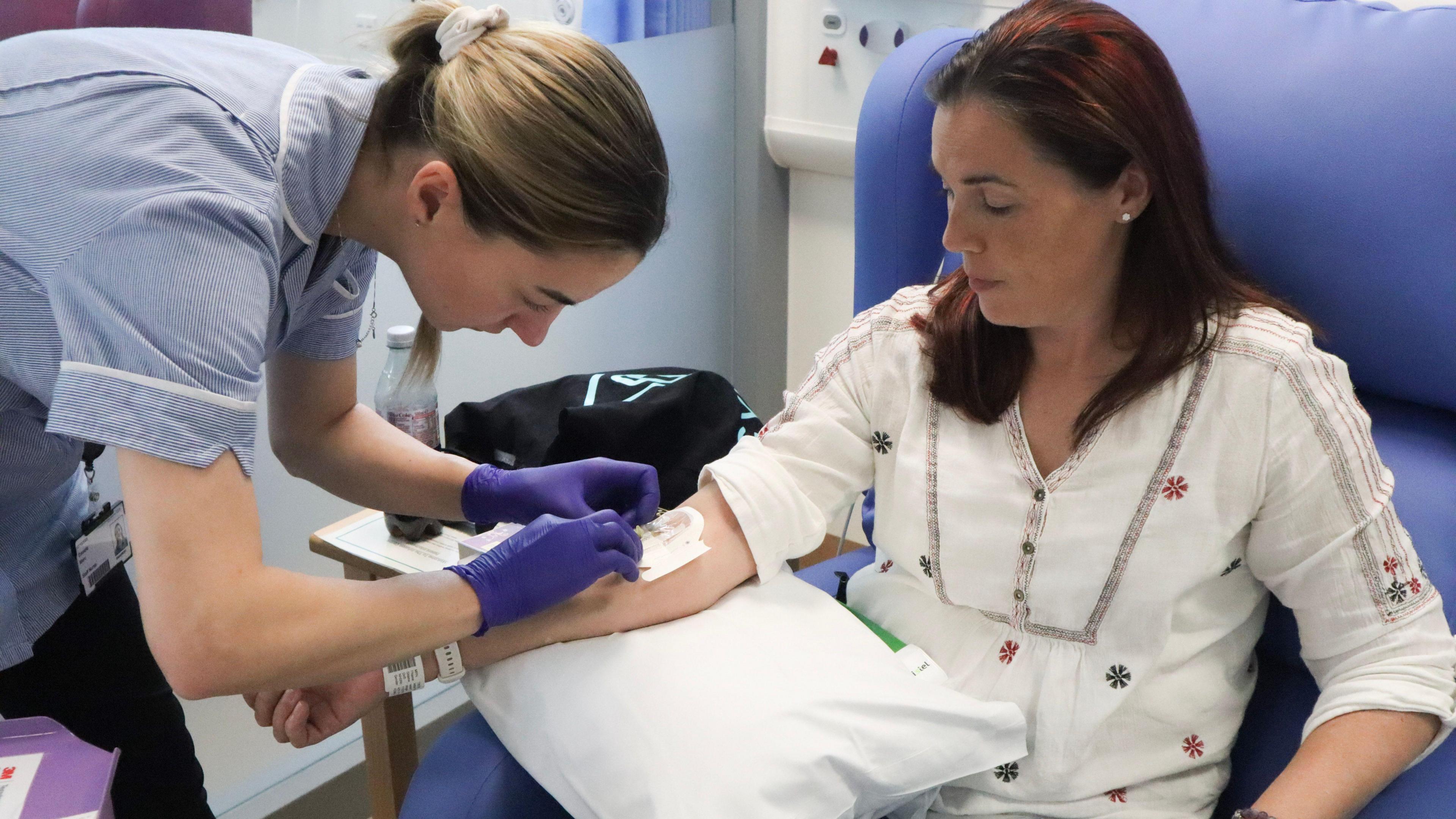 A healthcare professional in a blue uniform and purple gloves is administering treatment to a patient, Ms Wright, who is sitting in a medical chair. Her arm rests on a white pillow with medical equipment visible in the background. She is about to receive her regular Infliximab infusion at the Addenbrooke's Hospital Infusion Centre.