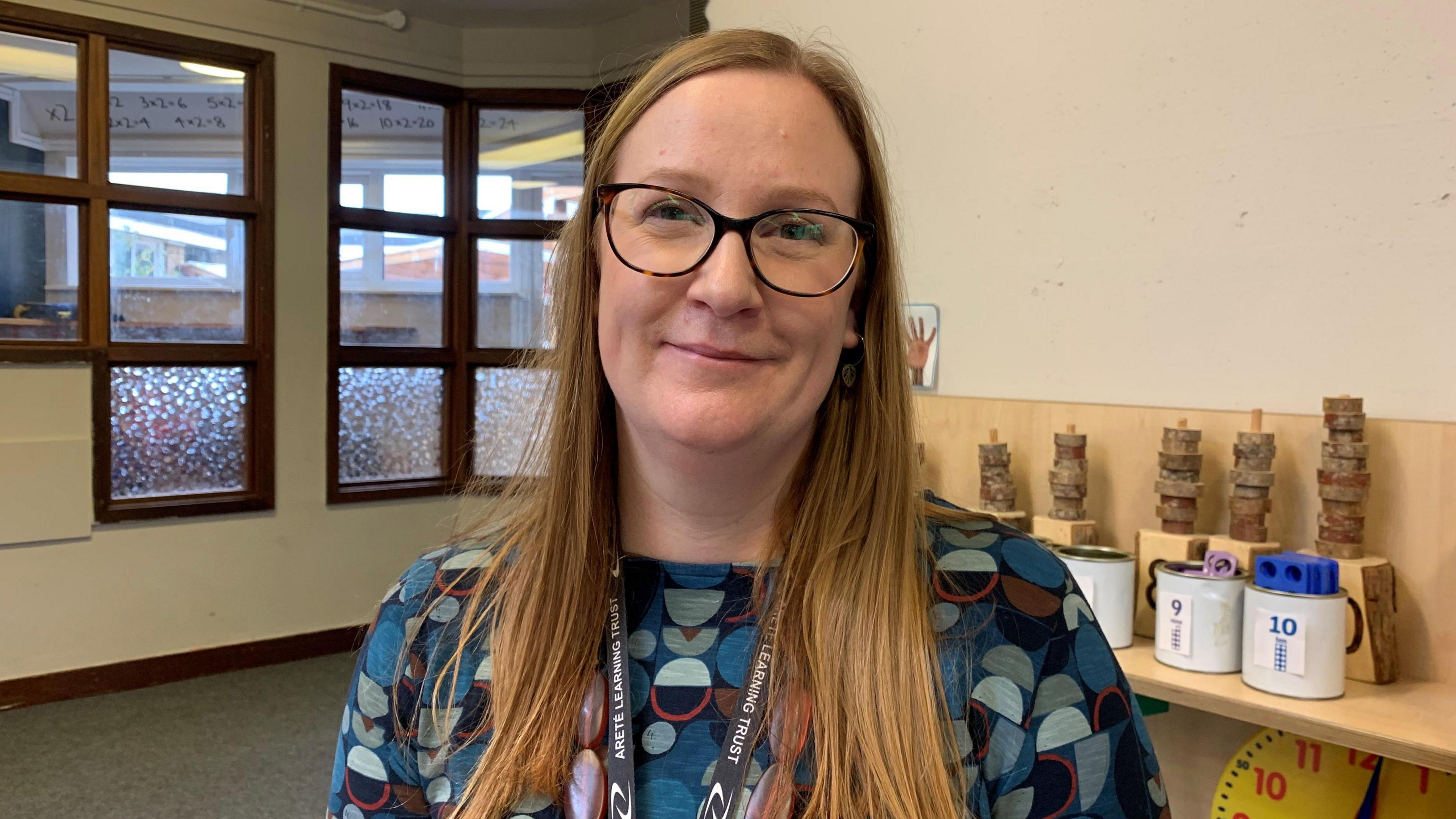 Kate Beck, an early years lead, wearing a white patterned shirt and dark blue v-neck jumper, pictured at a school library 