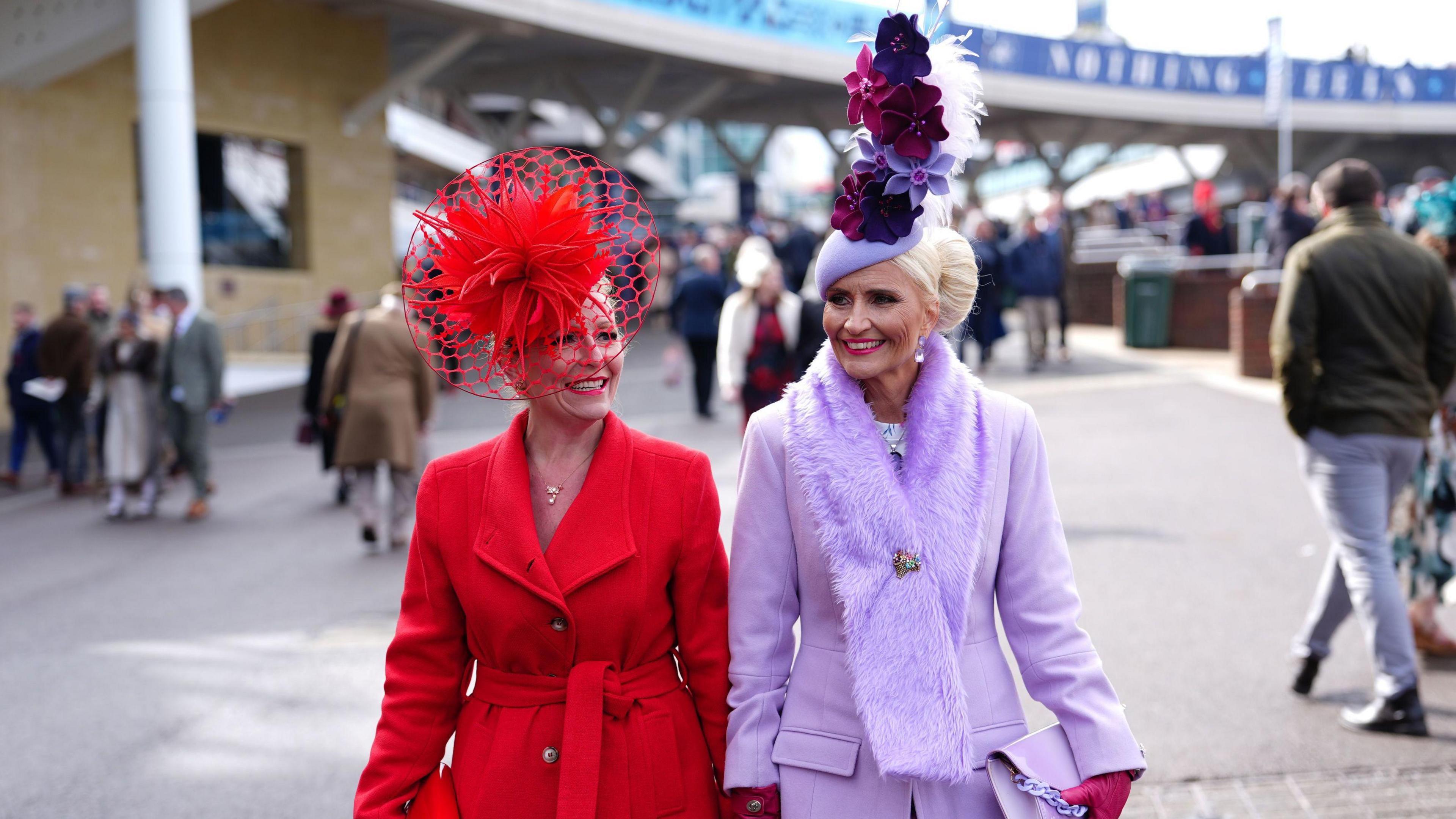 Two ladies wearing bright coloured outfits with large hats at Cheltenham Racecourse. 