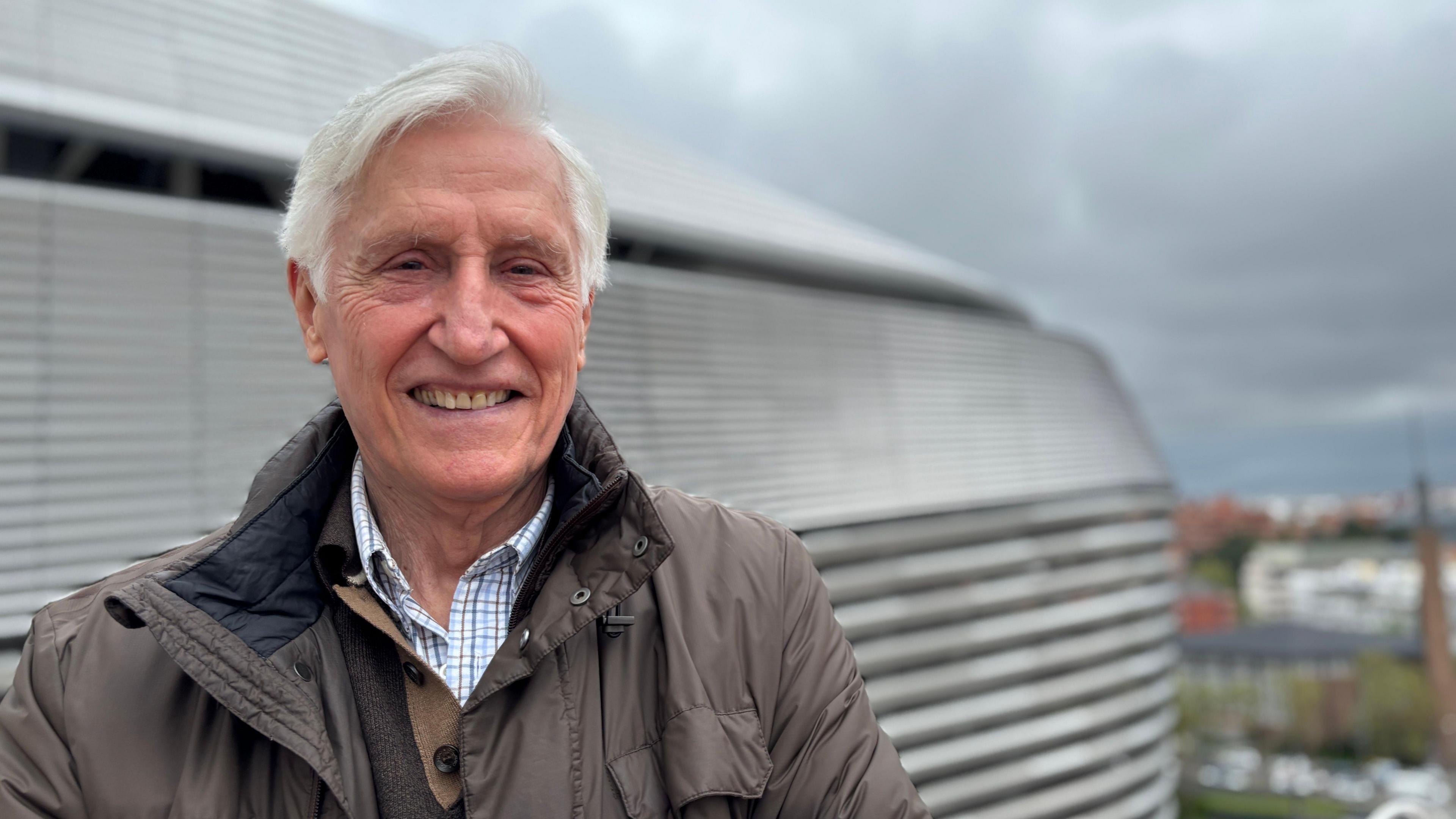 Elderly man with white hair slightly smiling towards the camera, with the Bernabeu stadium in the background.
