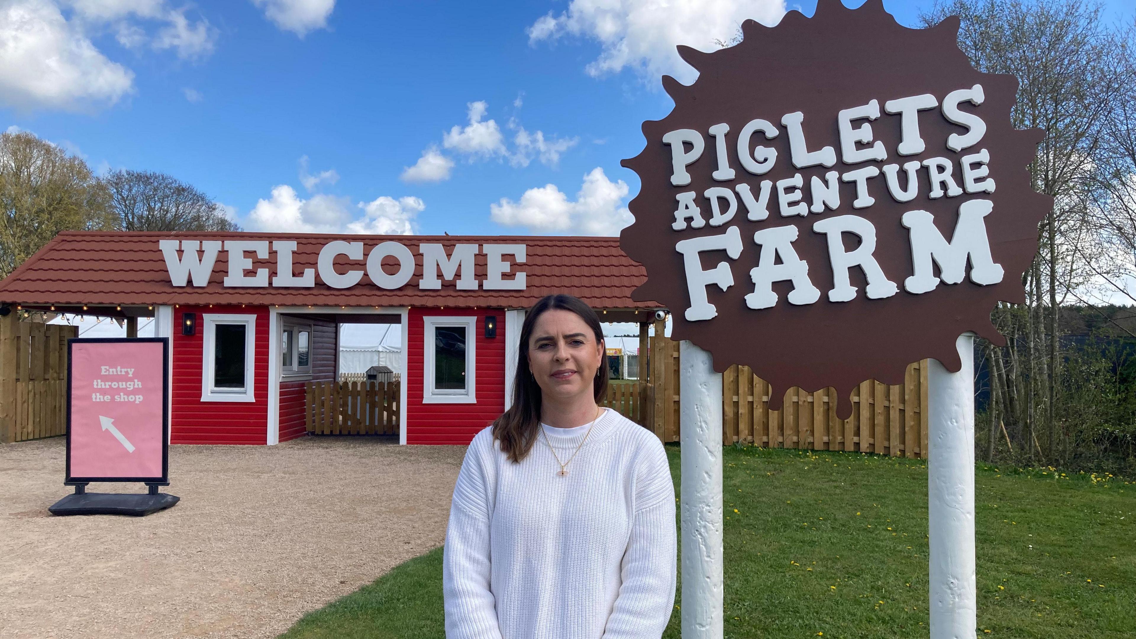An outdoor entrance area to a rural attraction.
Trees without dense foliage are visible in the background.
In the foreground, a woman is standing facing the camera. She is wearing a white long-sleeved sweater and a necklace.
A large, brown sign shaped like an irregular splatter reads in bold white capital letters: “PIGLETS ADVENTURE FARM”.
The sign is mounted on two vertical white posts fixed into the ground.
A smaller sign stands on the gravel path. It has a pink background and includes white text and an arrow. The visible text reads:
“Entry through the shop”
An arrow points inward toward the buildings.
Behind the person, there is a small red building with white trim and a brown tiled roof. The building has at least two small windows and resembles a ticket booth or reception hut.
Above the building entrance, large white capital letters spell:
“WELCOME”.