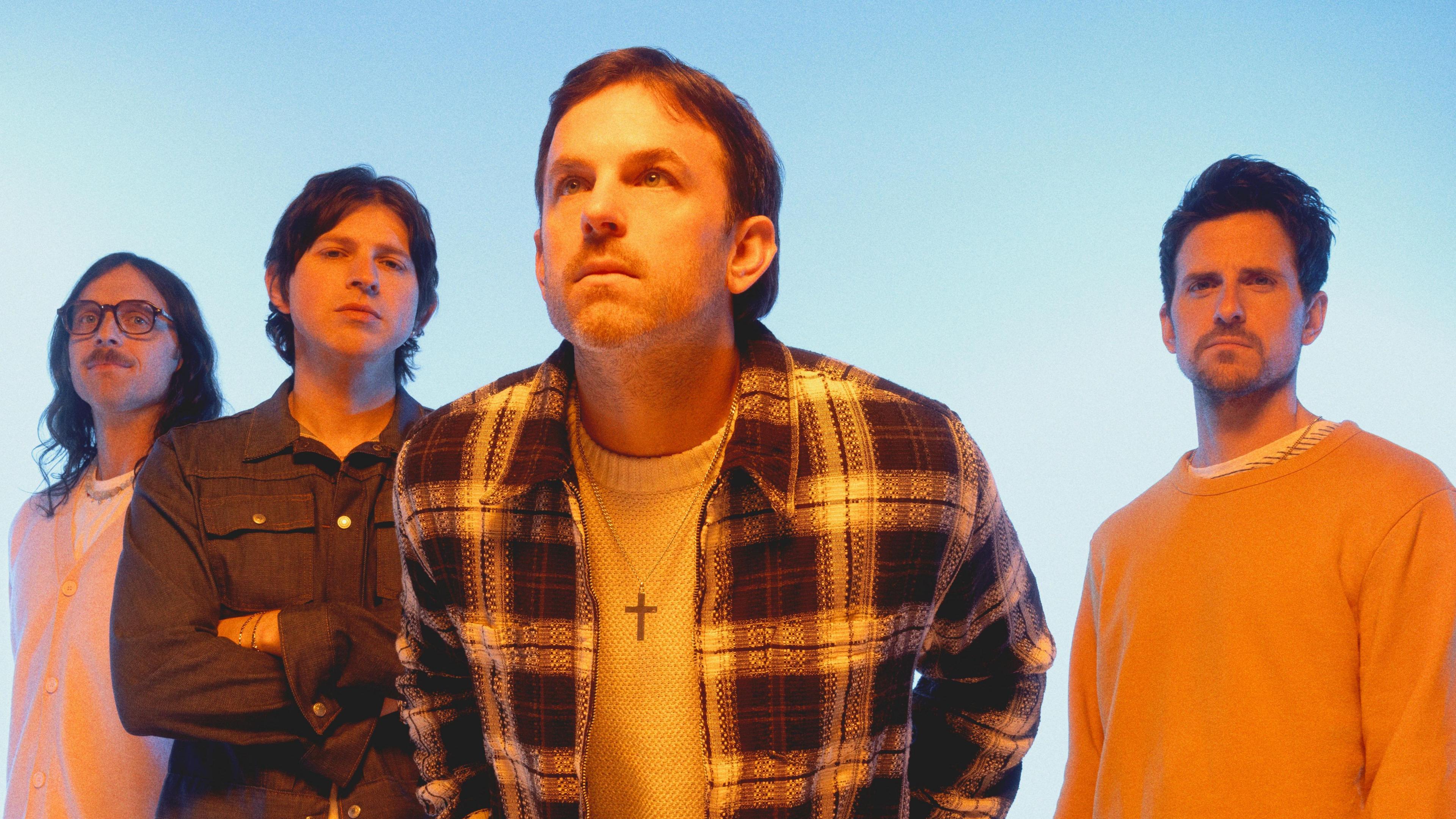 The four band members of Kings of Leon staring into the camera against a blue background. From left to right: Nathan Followill, who has long brown hair and a moustache, Matthew Followill, in a denim shirt, with his arms crossed. Cale Followil, leaning forward, with short brown hair and a crucifix around his neck, a checked shirt and a white top underneath. To his right, Jared Followill whose brown hair is swept back; he is wearing a yellow jumper.