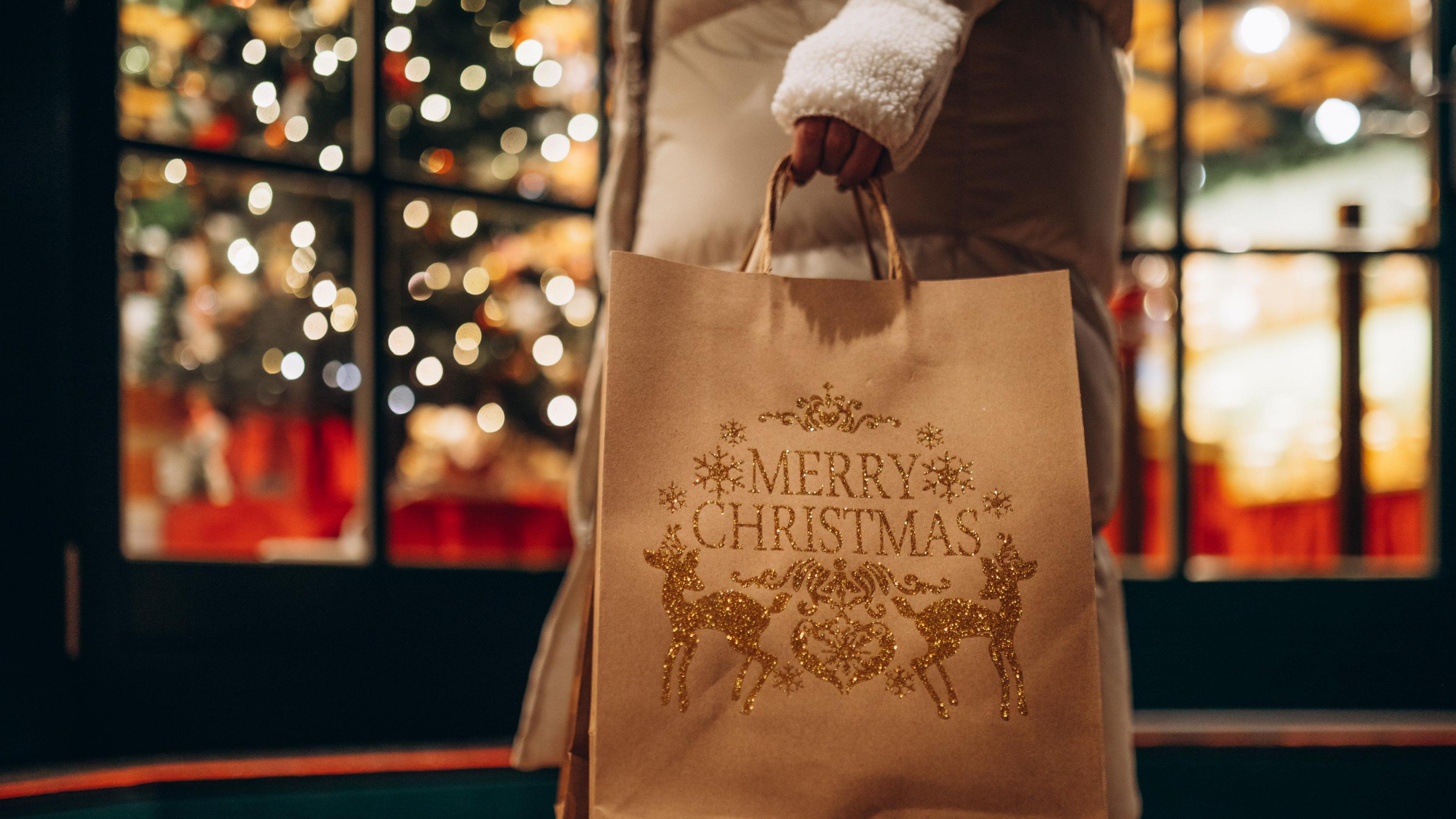 A woman wearing a brown coat with white fingerless gloves carries a bag decorated with "Merry Christmas" past a shop lit up for the festive season.